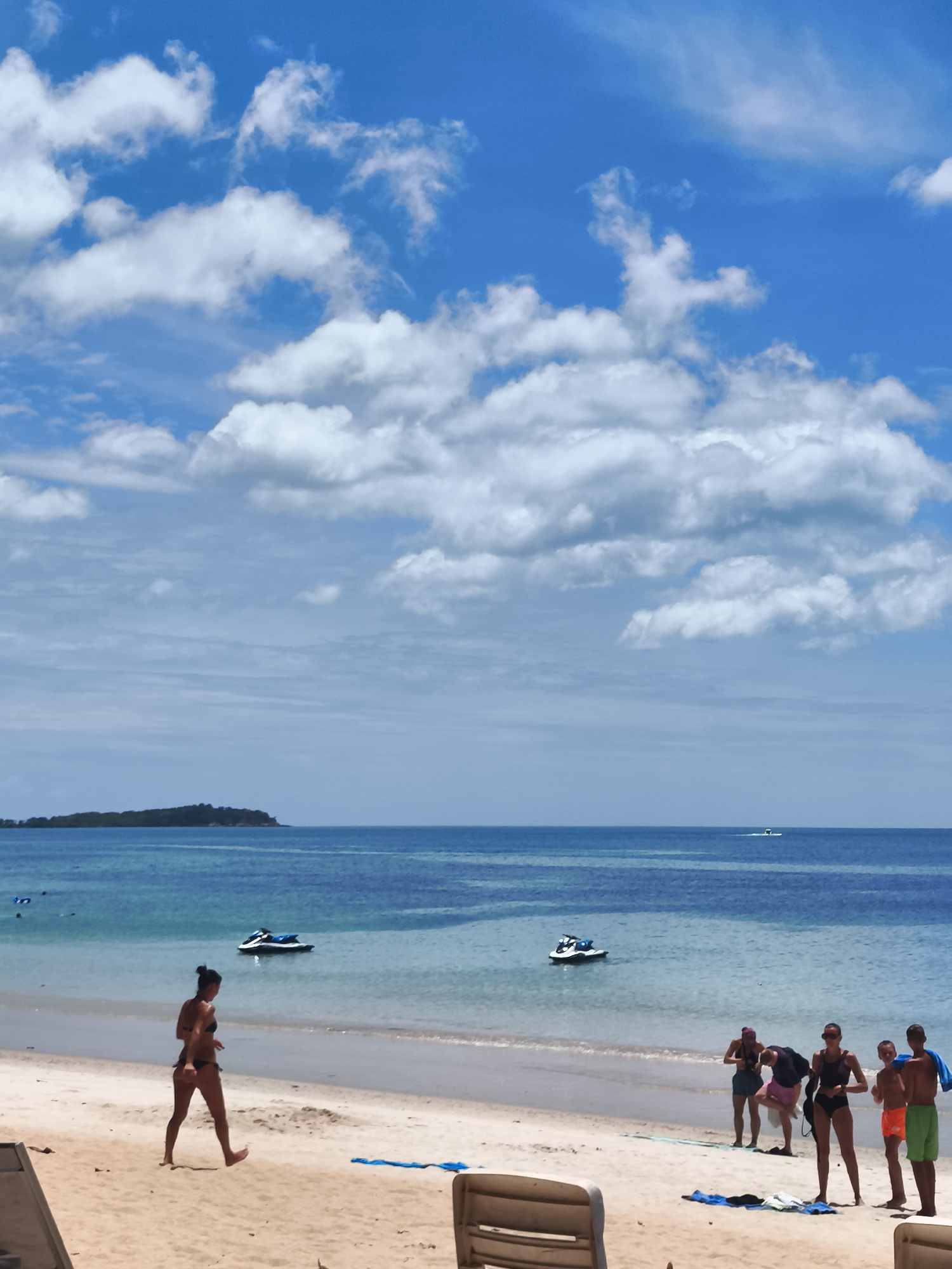 On a sunny day with a bright blue sky dotted with fluffy white clouds, a tropical beach scene unfolds in the Ko Samui District of Thailand. The turquoise waters of the sea gently lap against the sandy shore. In the background, a tree-covered island sits on the horizon.
Several people are enjoying the beach. A woman in a black bikini walks along the wet sand near the water's edge. Further down the beach, a group of people stand together, some appearing to be engaged in activities near the water. Two jet skis are visible in the shallow water, one closer to the shore and another further out. Beach chairs are positioned on the sand in the foreground. The overall impression is a relaxed and inviting atmosphere of a tropical vacation destination.