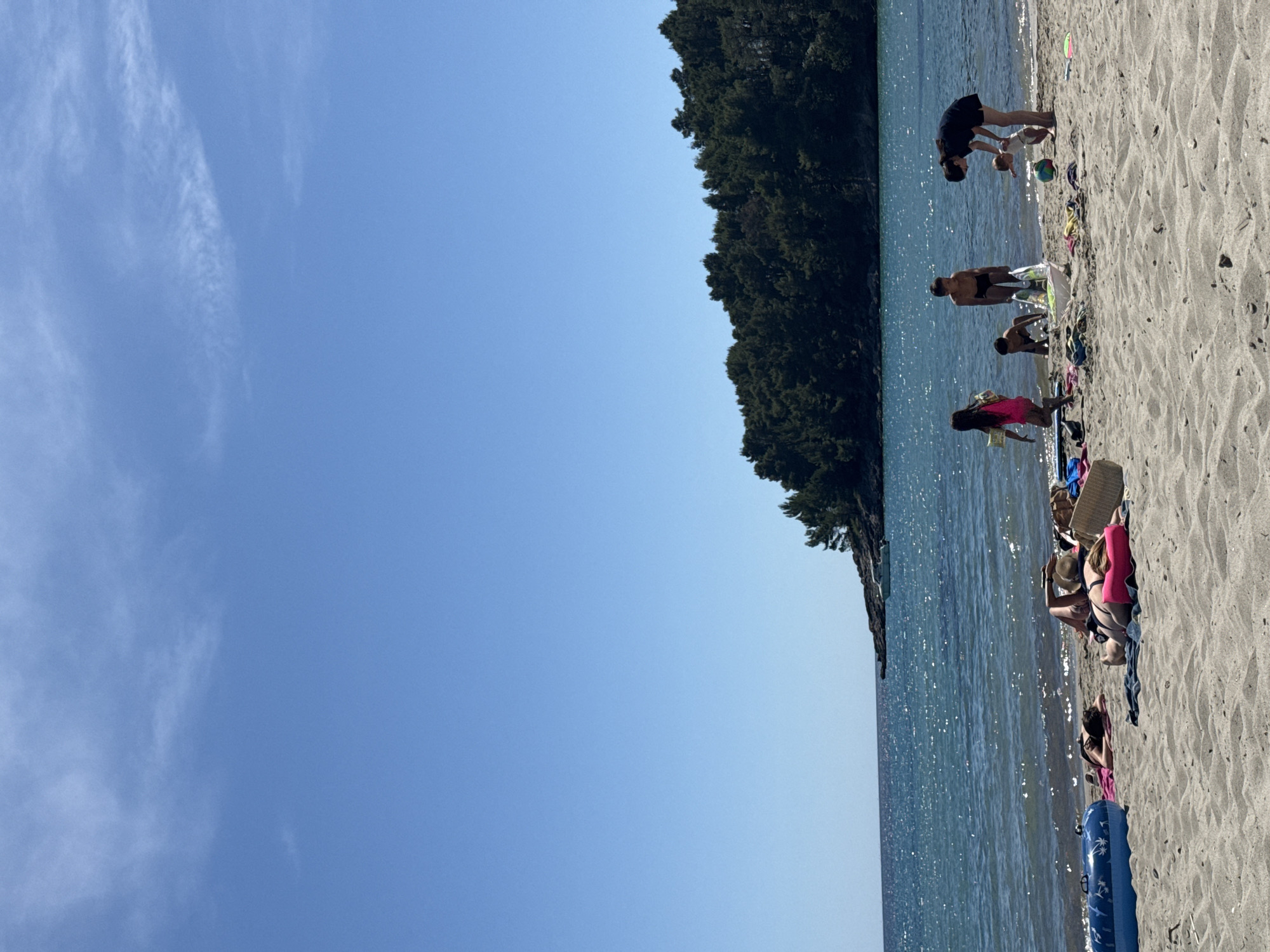 The image depicts a beach scene in Milovići, Montenegro on a sunny day. Several people are present on the sandy beach near the water's edge. Some individuals are standing, bending over, or appear to be playing with children, while others are sitting or reclining on the sand, likely sunbathing. The beach is adjacent to a body of water, potentially the Adriatic Sea, and a tree-covered area forms a natural boundary along the shoreline. The sky is clear with some clouds.