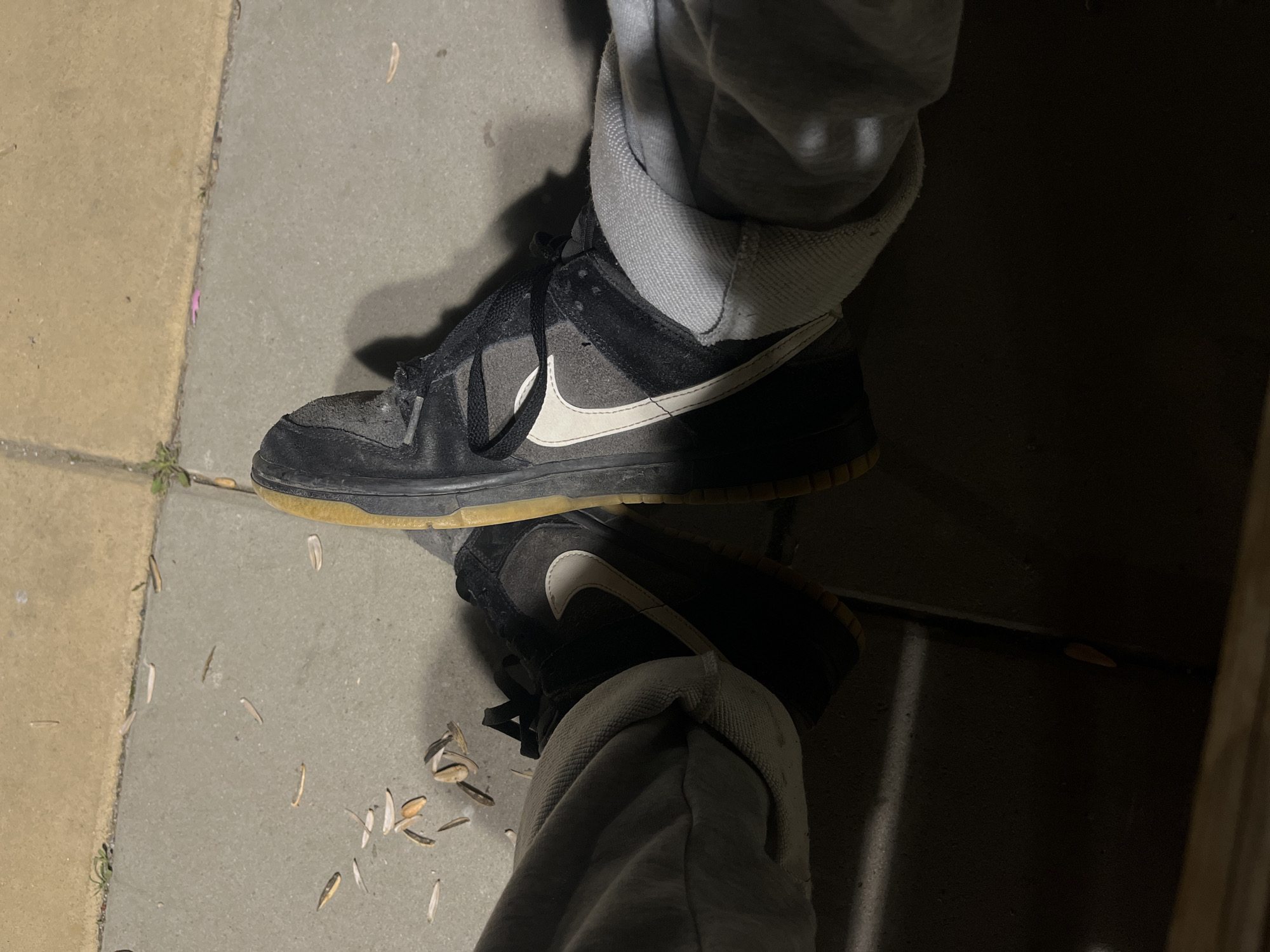The image depicts a person's legs and feet on a concrete surface, likely a sidewalk or walkway. The person is wearing gray sweatpants that are bunched around their ankles. Their shoes are black and gray Nike Dunk Low sneakers with a white swoosh and a yellowed sole. The shoes appear worn and have a distressed look. The ground around the feet is scattered with small debris, possibly flower petals or seed pods. The lighting is somewhat dim, suggesting it could be evening or the location is shaded. There are no specific details in the image to confirm it is Lozarevo, Bulgaria.