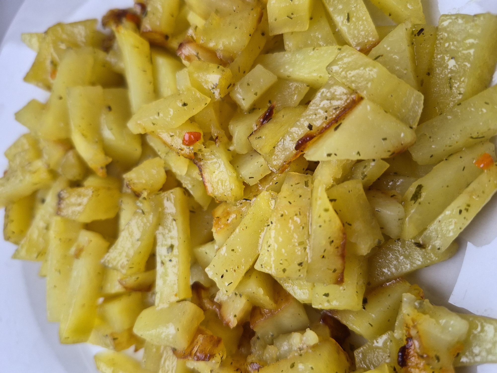A close-up photograph shows a pile of golden-yellow roasted potatoes. The potatoes are cut into various shapes, including small cubes and longer, rectangular pieces. They appear to be seasoned with herbs, evident from the small green specks distributed across their surfaces. Some pieces have slightly browned edges. A few small, red, pepper-like particles are visible, suggesting a spicy seasoning. The potatoes are arranged on a white plate. The image does not provide enough information to determine the location within Tobolsk, Russia, or any details about people, actions, interactions, or the broader environment.