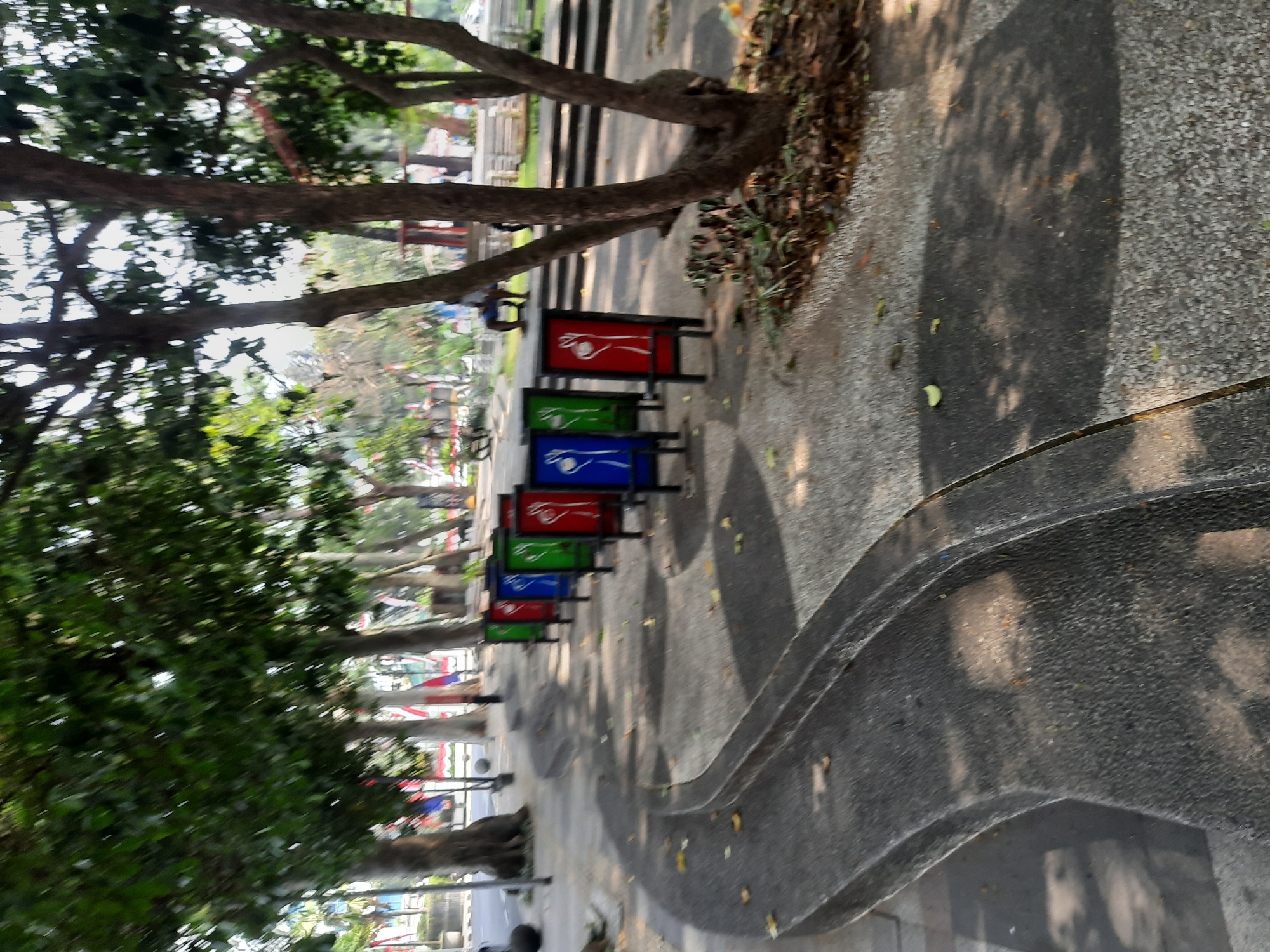 The image shows a park in Bandung City, Indonesia, with a curved concrete path in the foreground. Several colorful signs with white figures are lined up along the path. Trees with dense foliage are visible in the background, along with some buildings and flags. The lighting suggests it is daytime, with shadows cast on the ground. There are no people clearly visible in the image.