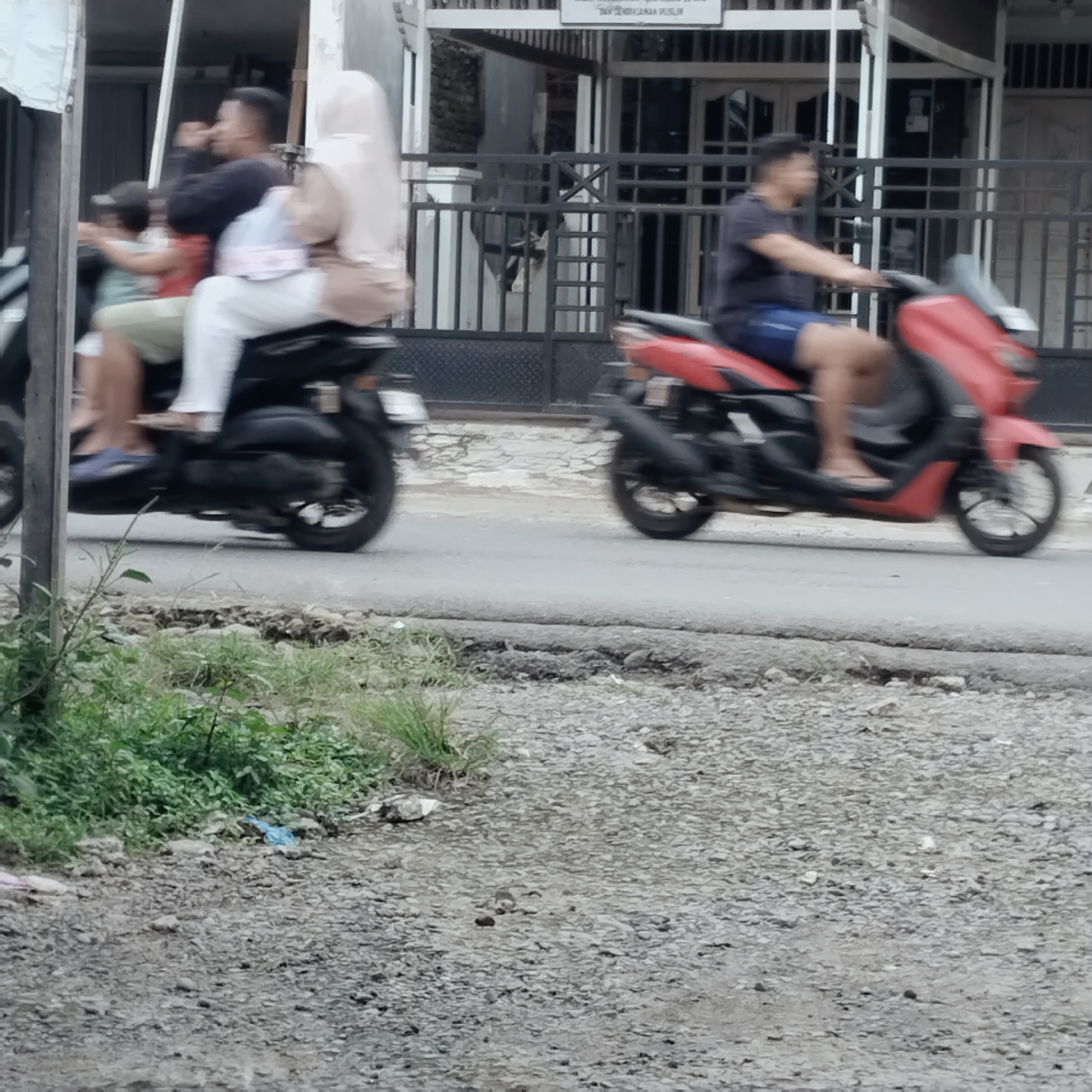 In Talun Kenas, Indonesia, two motorcycles are traveling on an asphalt road. The motorcycle on the left carries three individuals: a person driving, a person in the middle wearing a light-colored headscarf and white pants, and a child in the front wearing a green shirt and a cap. The motorcycle on the right is driven by a person wearing a dark shirt and blue shorts. Both motorcycles are in motion, indicated by the blur of the wheels and the road. The environment appears to be a roadside with gravel and vegetation on the left side of the frame. Buildings with barred windows and a fence are visible in the background.