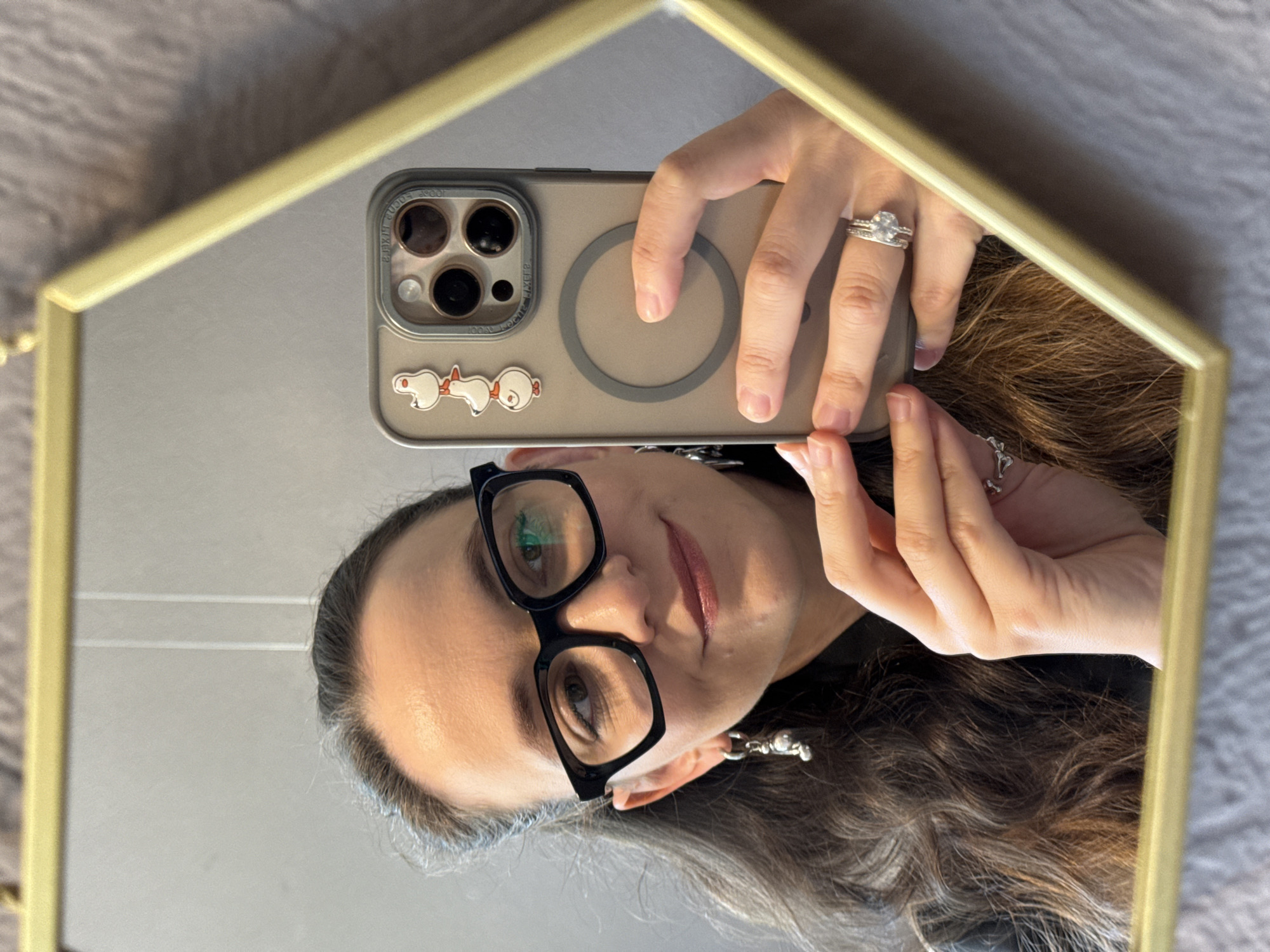 A young woman is taking a selfie in a hexagonal mirror. She has long, wavy brown hair, is wearing black-rimmed glasses, and has on a black top. She is also wearing a silver ring on her left hand, a ring with a large stone on her right ring finger, and a pair of silver earrings. She is holding her phone, which has three duck stickers on it and a clear case. The background of the image shows a wall and a textured surface. There is no indication of the city of Chaikovsky, Russia in the image.
