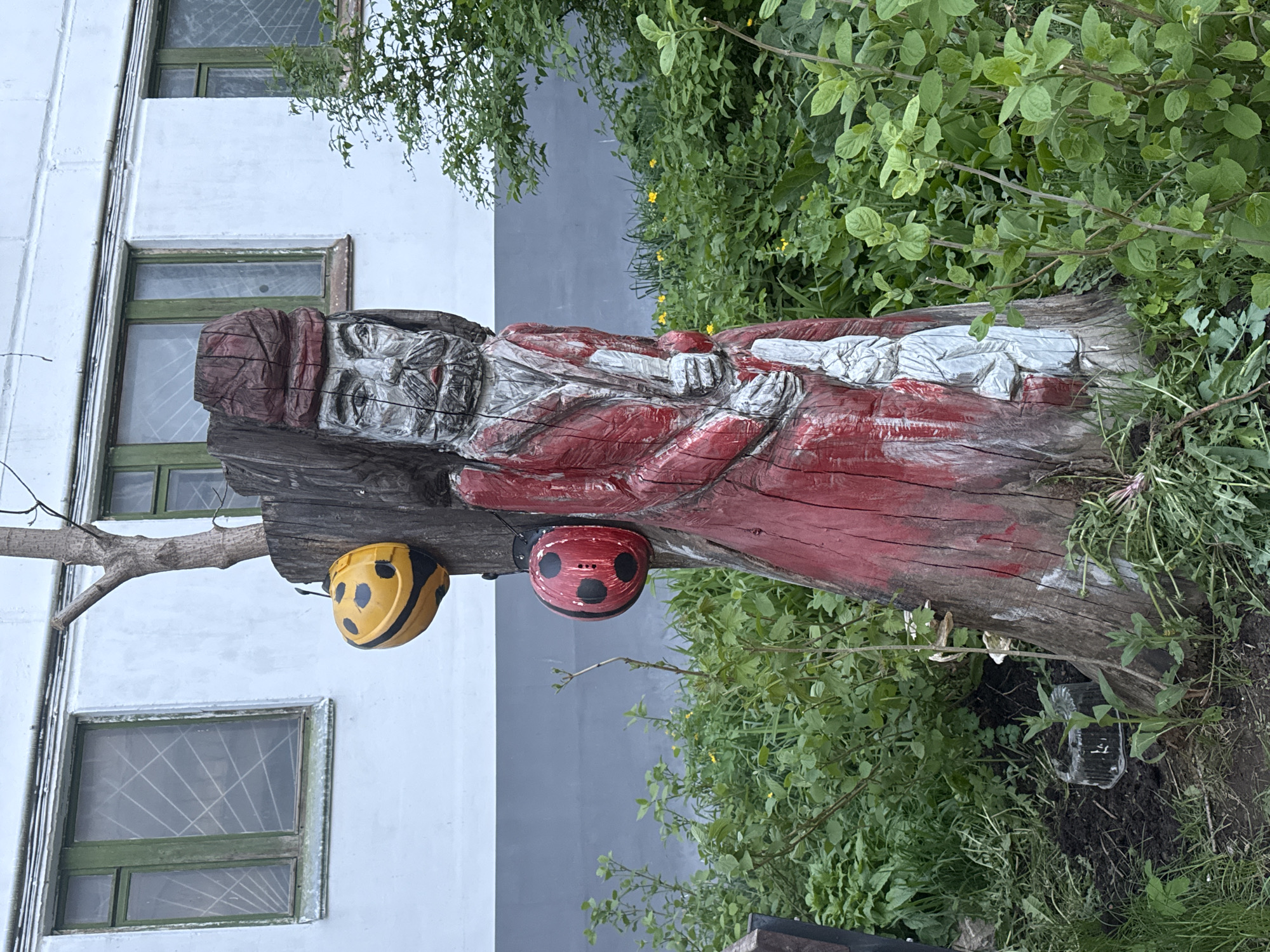 The image depicts a carved wooden statue of a bearded figure wearing a red garment. The statue is located outdoors amidst greenery, with a light grey building in the background. Two ladybug-like spherical decorations, one yellow and one red, are attached to the wood near the statue's head. The building has two windows with green frames and a wire mesh over the glass. The statue is likely located in Chaikovsky, Russia.