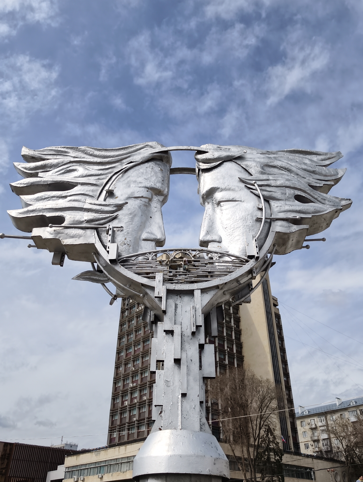 The image shows the "Zhuravli" (Cranes) monument in Saratov, Russia. This modern sculpture features two stylized, silver-colored metal faces, each with flowing hair, positioned to look at each other within a circular frame. The frame is supported by a tall, metal structure with geometric shapes and a wide base. The monument is placed outdoors in an urban environment. Behind the sculpture are buildings, including a tall building with multiple rows of windows, suggesting it's an office or apartment building. The sky is visible in the background, displaying a mix of blue and light, scattered clouds. Bare trees are also visible to the right.