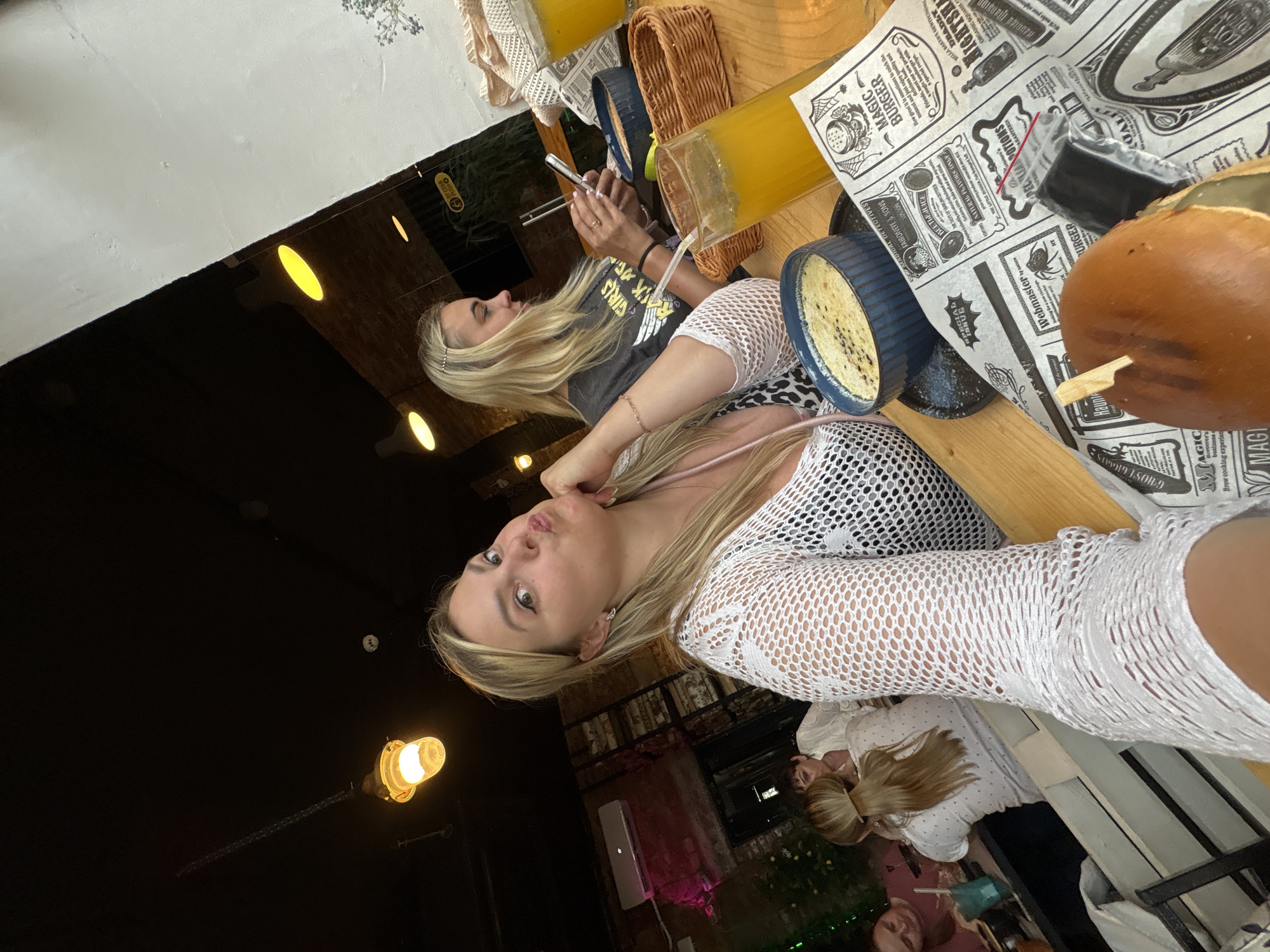 Two young women with blonde hair are seated at a wooden table, seemingly at an outdoor restaurant or cafe. The woman in the foreground is looking directly at the camera, resting her chin on her hand and making a pouty face. She is wearing a white, lacy top. The second woman, further back, is looking down at her phone, which she is holding with chopsticks. She has on a dark t-shirt with a graphic design.
On the table, several objects are visible, including a glass of orange juice, a woven basket, and two blue bowls containing food that appears to be a savory dish with speckles. There are also pieces of paper spread out on the table, which seem to be themed menus or placemats with text like "MAGIC BURGER," "Haunted Mansion," and "DEADLY NIGHTSHADE." The lighting suggests it is evening, with warm yellow lights from overhead fixtures creating an ambient glow. The overall mood seems casual and relaxed, with the women enjoying their food and drinks. The scene takes place in Chaikovsky, Russia.