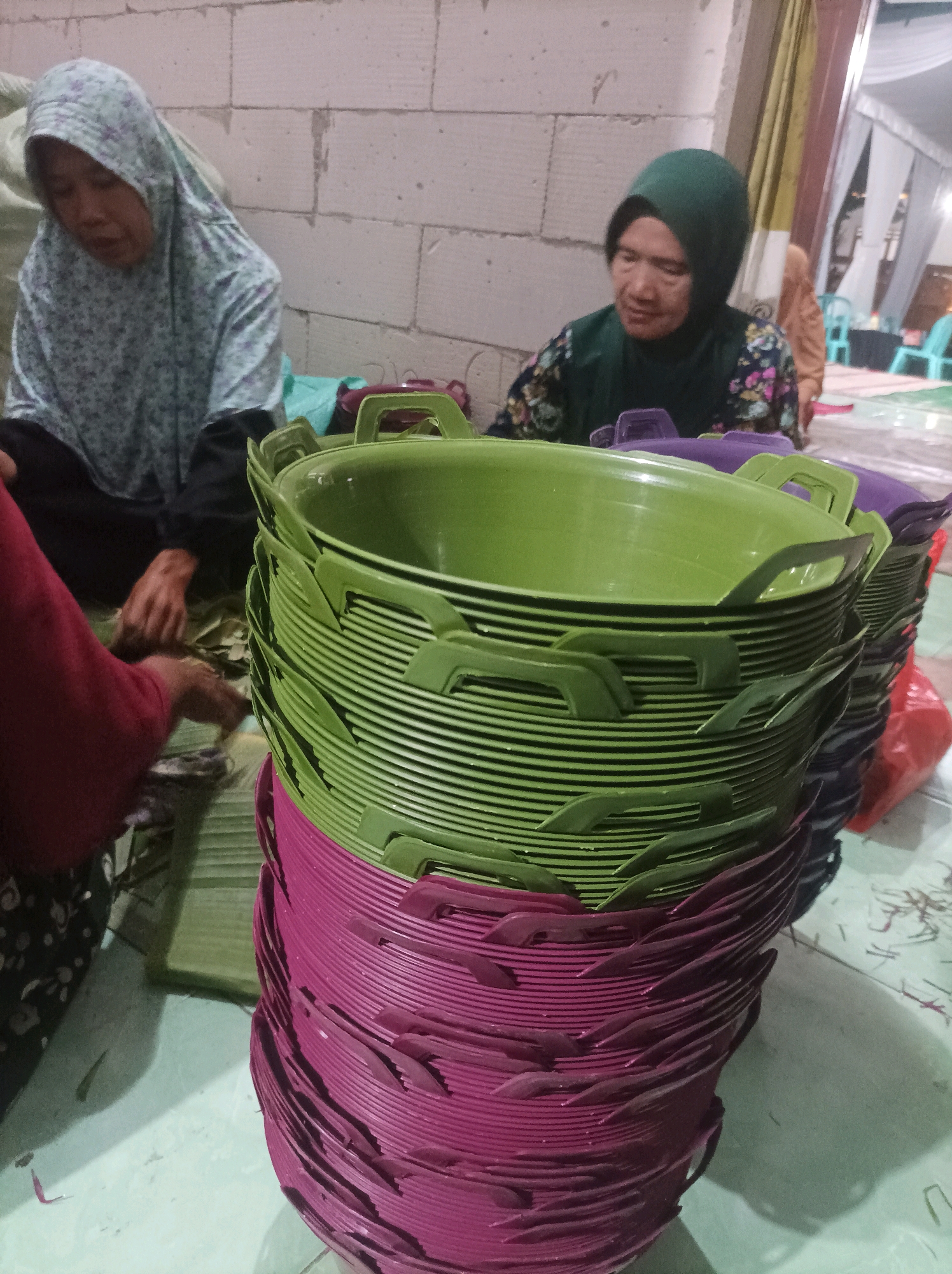 In a dimly lit indoor setting, likely a residence or workshop in Canga'an, Indonesia, two women are engaged in a task. One woman, wearing a floral-patterned hijab, is seated on the floor in the foreground on the left, her hands occupied with what appears to be arranging banana leaves. She is wearing a dark-colored top and black pants. To her right, a large stack of brightly colored plastic baskets dominates the center of the frame. These baskets are a vibrant green at the top, transitioning to a deep purple at the bottom. They are stacked in neat rows, with their handles protruding outwards.
Behind the baskets, another woman is seated, wearing a green hijab and a dark floral patterned blouse. She is looking down and appears to be working on something, though her actions are less clear than the woman in the foreground. In the background, a wall constructed of white cinder blocks is visible. To the far right, a white tent or canopy structure is partially visible, along with glimpses of turquoise plastic chairs, suggesting a communal gathering or event might be taking place or has recently concluded. The floor is a light-colored tiled surface. The overall scene depicts a domestic or small-scale production environment.