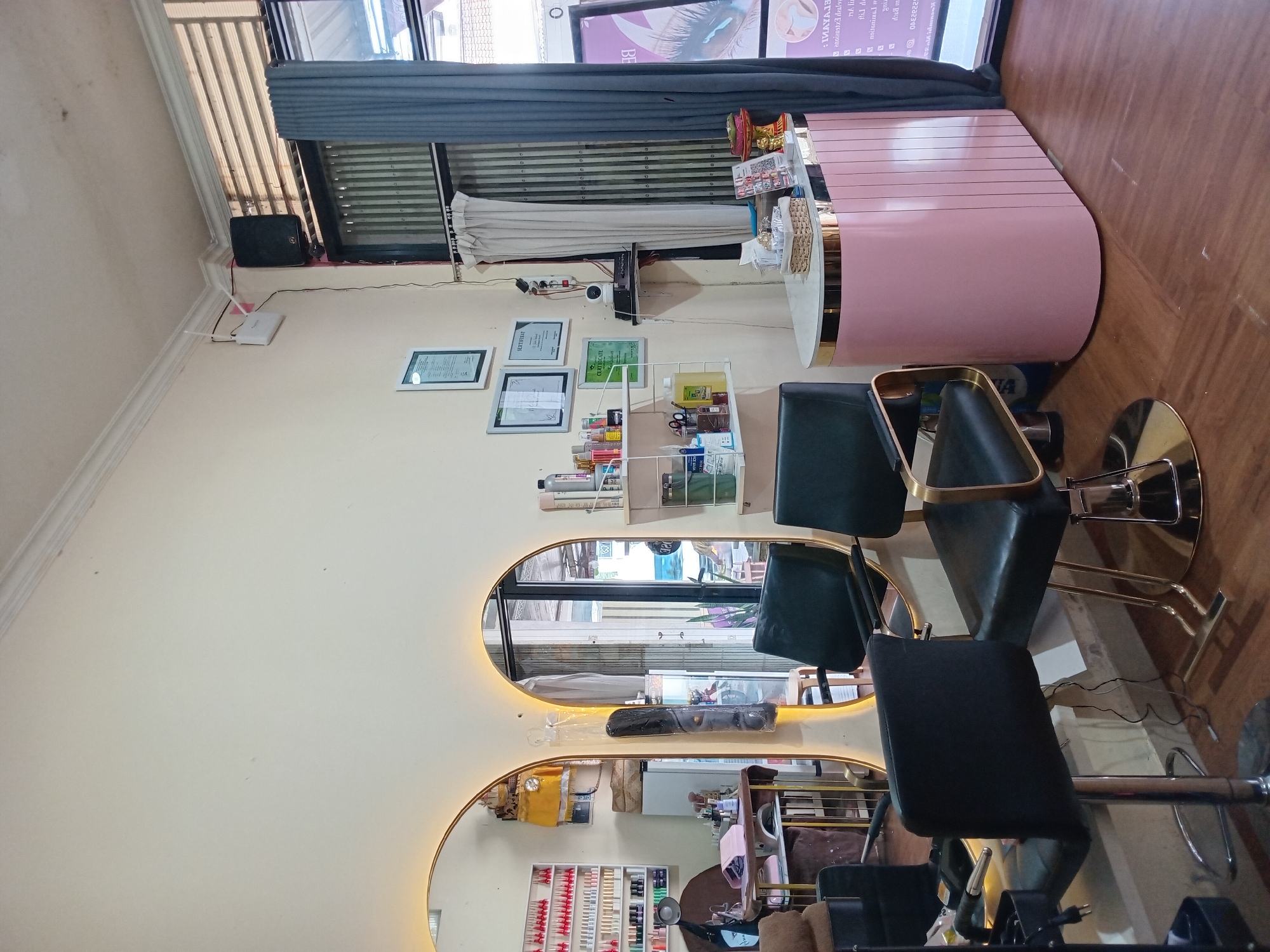 This image captures the interior of a beauty salon or spa located in North Kuta, Indonesia. The space is well-lit and appears ready for customers, though no people are present.
The focal point is a modern pink and gold reception desk situated on the right, with a small decorative item on its surface. In front of the desk are several black and gold hydraulic salon chairs, suggesting areas for services like hair styling, manicures, or pedicures. The floor is made of polished wood.
Along the left wall, there are three framed certificates, with text including "CERTIFICATE" and "SERTIFIKAT," indicating professional qualifications or achievements. Below these, a white shelf holds various beauty products in bottles, jars, and other containers. Further along the wall are two large, vertically aligned oval mirrors, each featuring integrated backlighting that casts a warm glow. Reflected in the mirrors are views of the opposite wall and additional salon equipment, including what looks like a nail polish display.
Near the ceiling on the left, a white router/modem with an antenna is mounted, next to a black wallet and beneath a window fitted with horizontal blinds and drapes. Another set of curtains hangs in the background near a window displaying mirrored text.
Visible text on the window reflection includes "BE", ": IMAYA13", "anoianstza delova" (which appears to be mirrored text for a beauty service like "LASH EXTENSION"), and "2202340" (possibly a contact number or ID). The overall setting is clean, modern, and professional, characteristic of a contemporary beauty service establishment.
