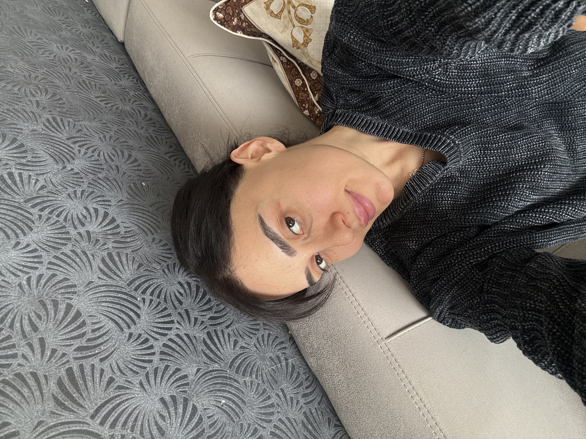 An indoor, slightly rotated, eye-level shot captures a woman relaxing on a light beige sofa, likely in Tbilisi, Georgia. The woman is positioned on her side, with her dark hair falling around her head. She has dark, well-defined eyebrows, brown eyes, and a fair complexion. Her head is turned towards the camera, and she looks directly at the viewer with a soft, engaging smile. She is wearing a dark gray or black knitted sweater with a visible textured pattern.
A decorative pillow with a brown and beige floral pattern is partially visible behind her head on the sofa. The sofa itself is upholstered in a plain, light beige or light gray fabric, featuring distinct stitched seams along its cushions. The floor to the left of the sofa is covered by a dark gray rug or carpet, which displays an intricate, swirling pattern of lighter gray or silver fan-like motifs. The lighting appears bright and even, indicating an indoor setting with ample illumination. The woman's relaxed posture and gentle expression convey a sense of calm and comfort. No text is visible in the image.