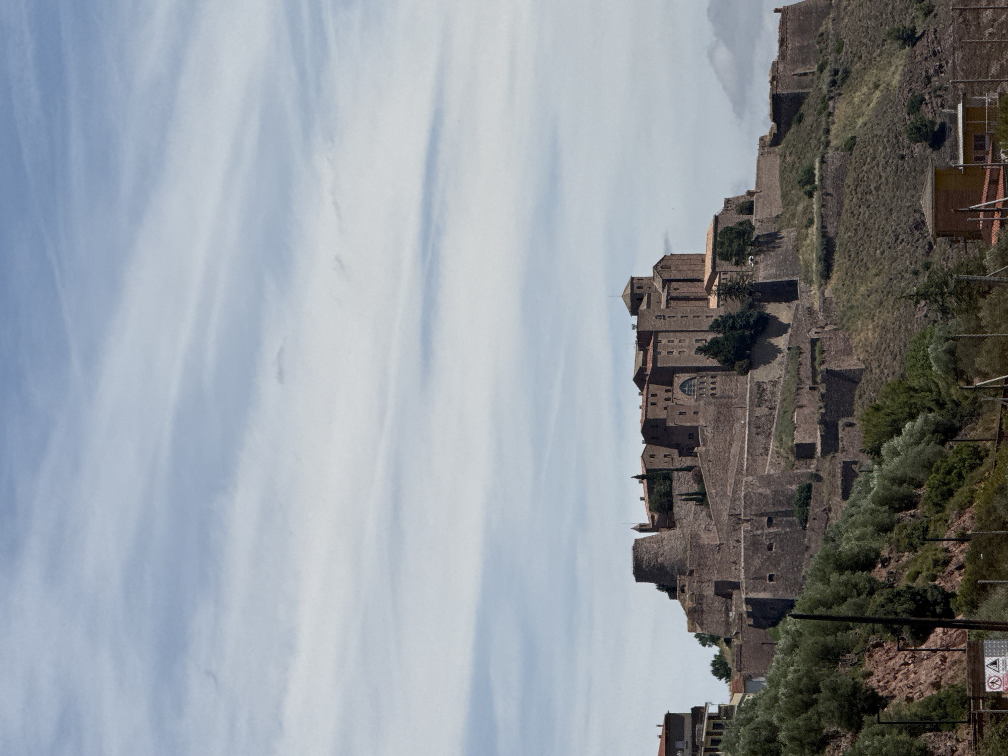 The image shows a medieval castle situated atop a hill overlooking a valley. The castle is constructed of stone and features multiple towers and defensive walls. Lush green vegetation covers the slopes of the hill, with olive trees interspersed among the rocky terrain. The sky above is a clear blue with wispy white clouds. The castle appears to be well-preserved and is a prominent landmark in the city of Cardona, Spain. In the foreground, there are some modern structures, including a fence and a yellow building, which suggest the presence of contemporary development near the historical site. No people are visible in the image.
