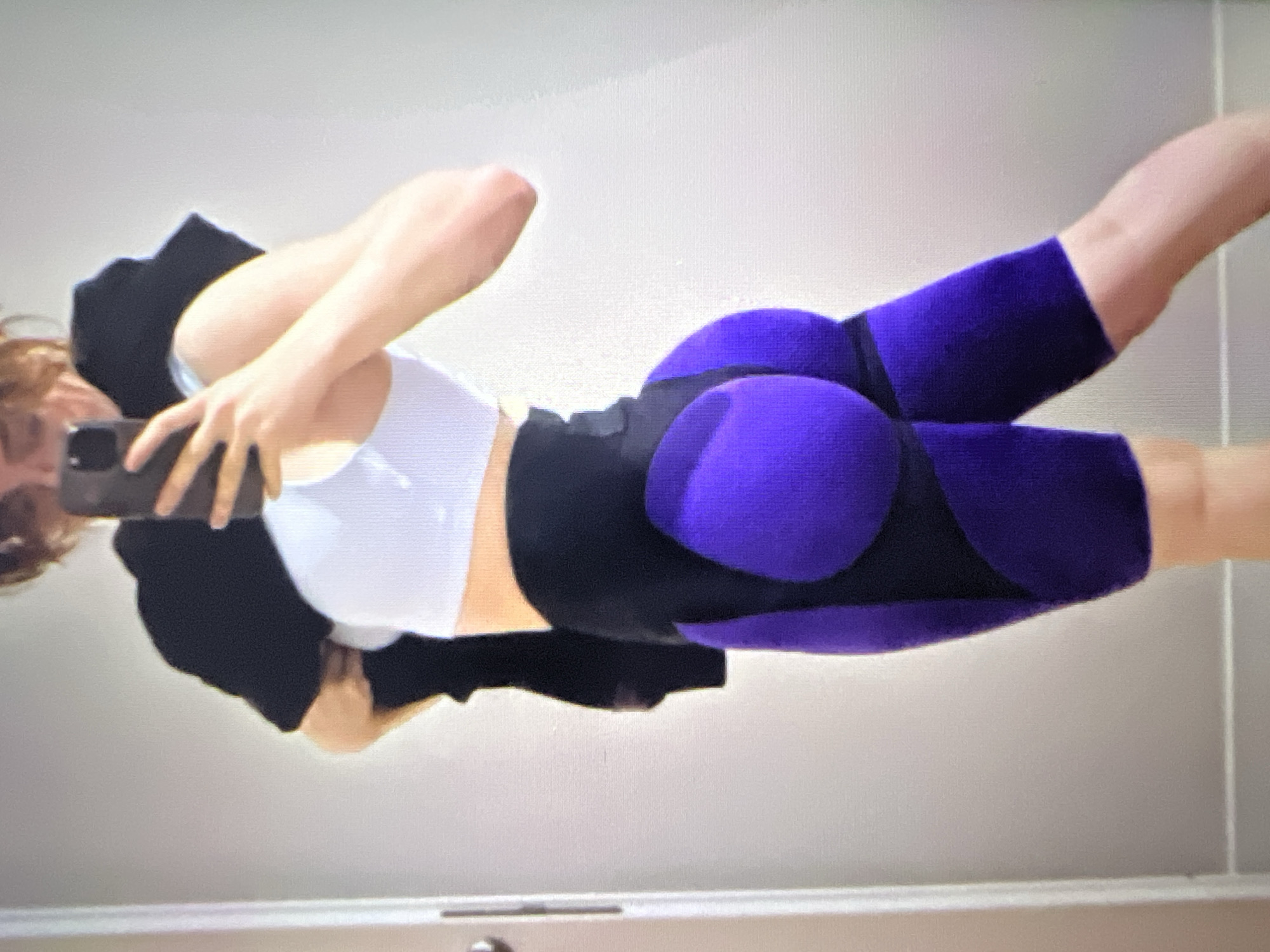A person is standing in a room, taking a mirror selfie with a smartphone. They are wearing a white sports bra, black shorts with purple accents that emphasize the buttocks, and a black hooded sweatshirt draped over their shoulders. The background appears to be a wall with a door frame on the left. The lighting is bright, casting minimal shadows.