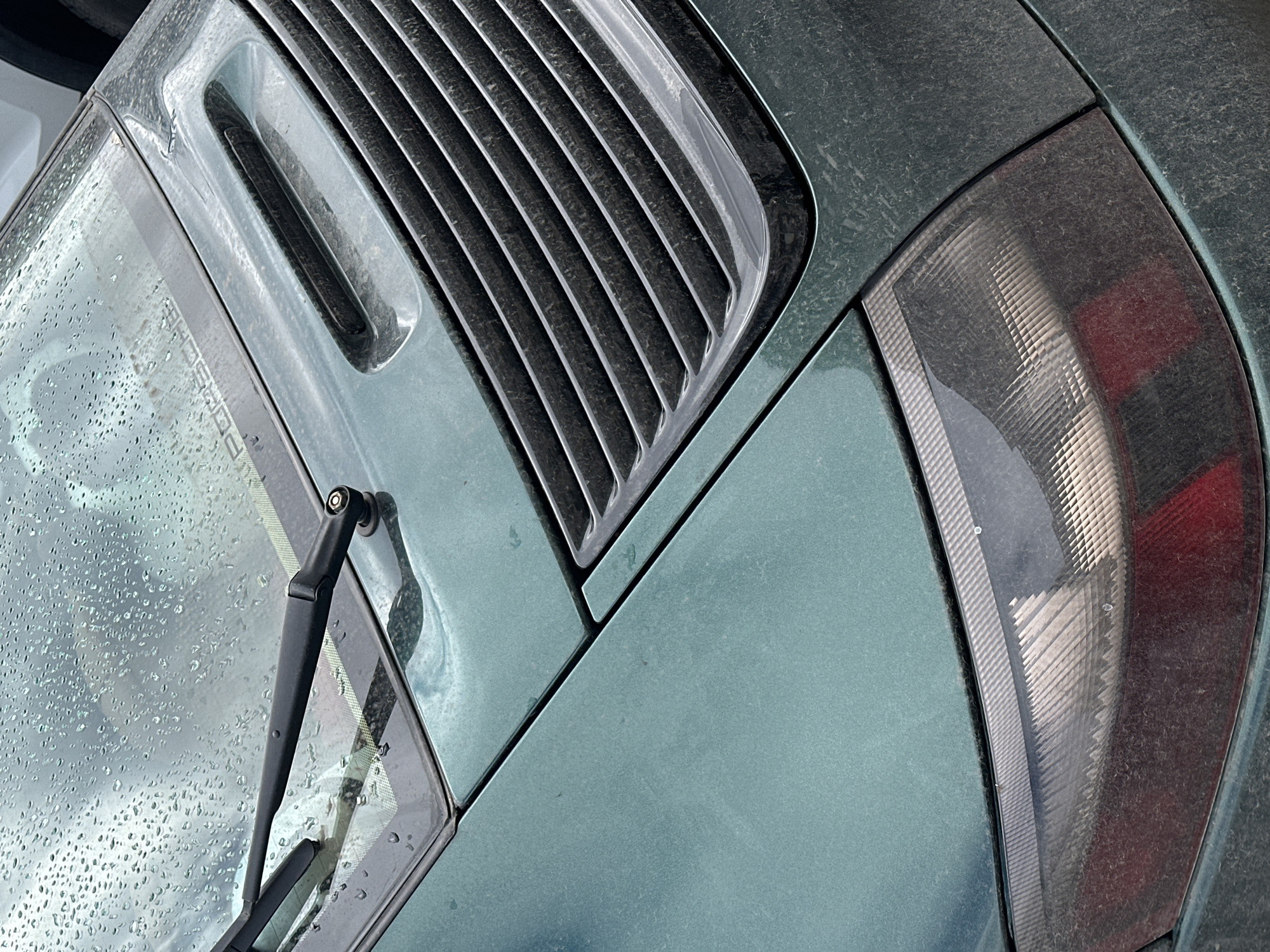 The image shows a close-up of the rear section of a dusty, possibly dark green car. Visible features include a rear window wiper, a series of vent slats, and a taillight. The car's surface is covered in dust or dirt. The location is not identifiable in the image.