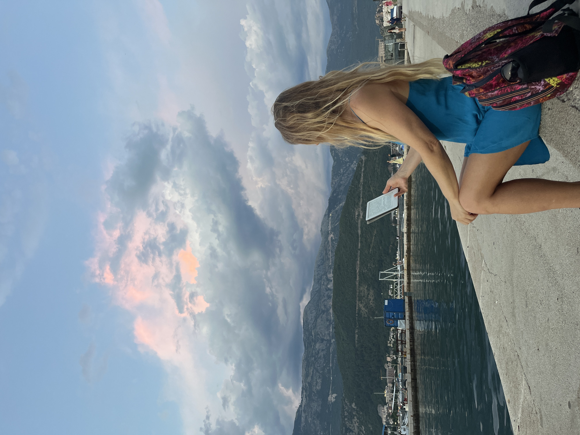 A woman with long blonde hair is sitting on a concrete promenade, reading an e-reader. She is wearing a blue dress and has a colorful backpack next to her. The scene is set in Budva, Montenegro, with the Adriatic Sea to her left and mountains in the background. The sky is filled with dramatic clouds, tinged with pink and orange from the setting sun. In the distance, there are boats docked in the harbor and buildings along the coastline.
