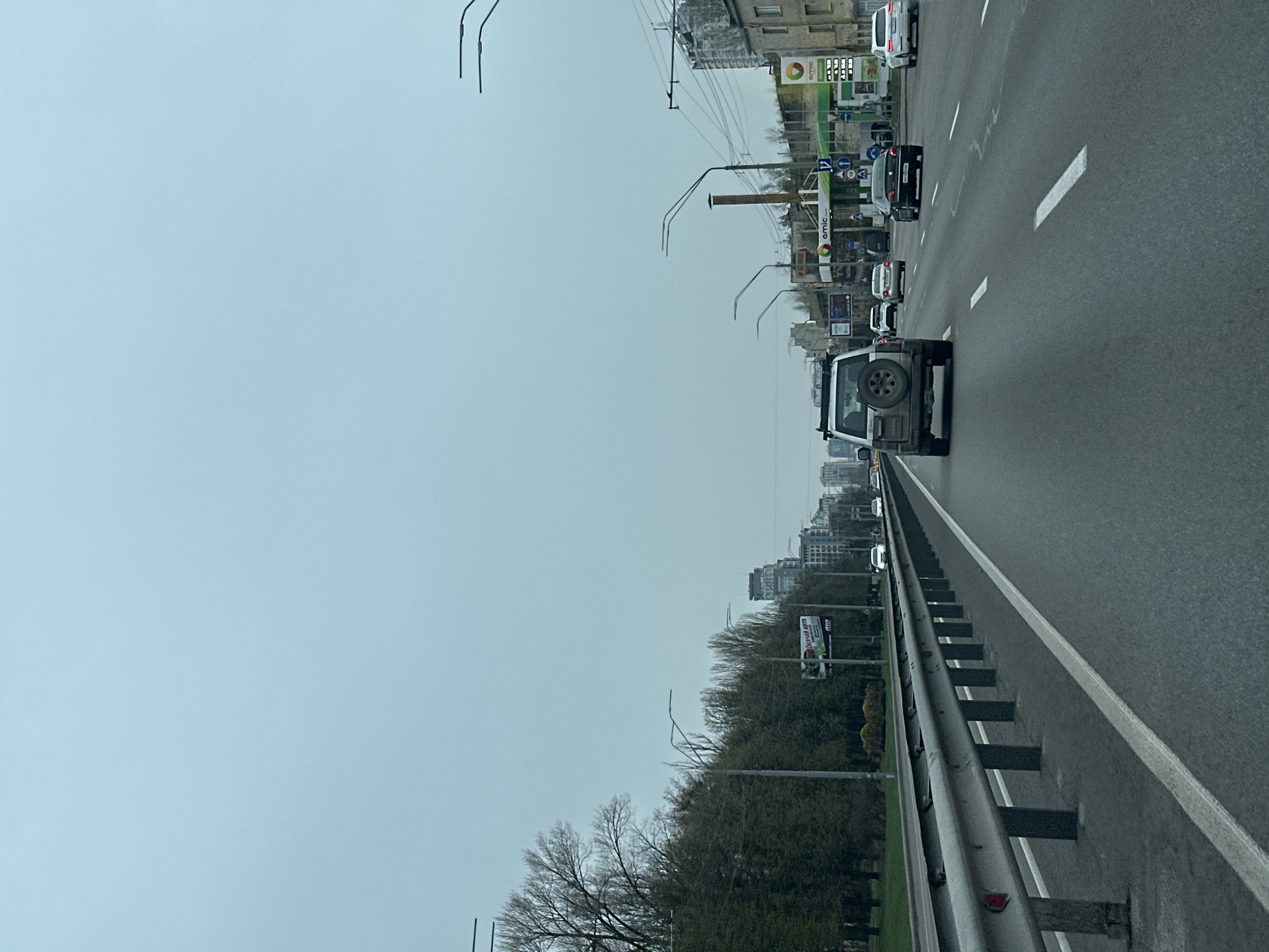The image depicts a view from a car traveling on a highway in Kyiv, Ukraine. The highway has multiple lanes, with cars traveling in the same direction. There is a guardrail on the right side of the highway. The skyline of Kyiv is visible in the distance on both sides of the highway. An AMIC gas station is visible on one side. A billboard is visible on the right side of the highway. The sky is overcast.