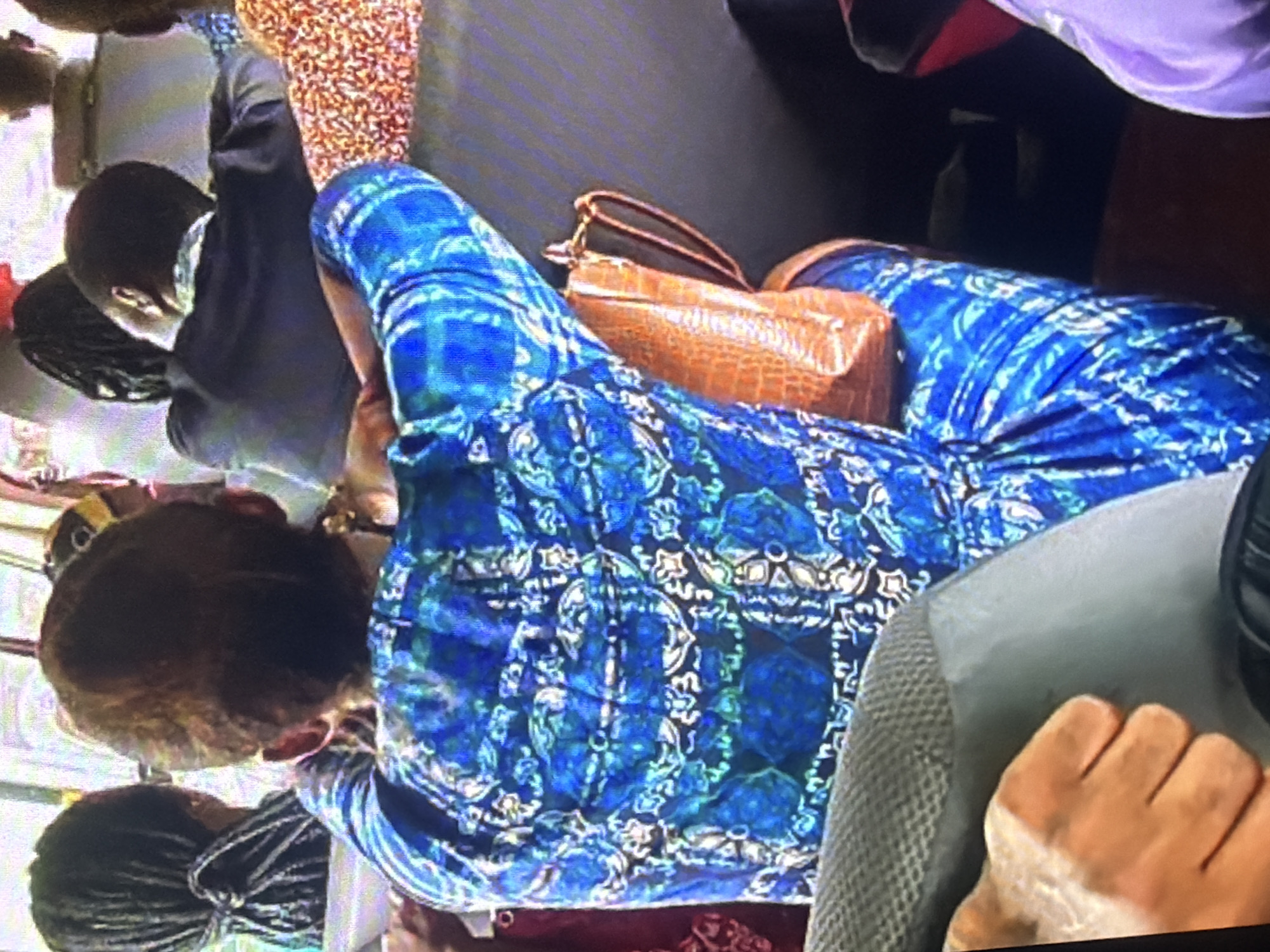 The image shows the interior of a bus with several passengers. In the foreground, a person wearing a blue and white patterned dress is seated, with their back to the camera. To their left, another person's hand rests on a grey seat. To the right of the person in the blue dress, a tan, textured handbag is visible. In the background, other passengers are seen from behind. The environment suggests a mode of public transportation within the city of Bukuru, Nigeria.