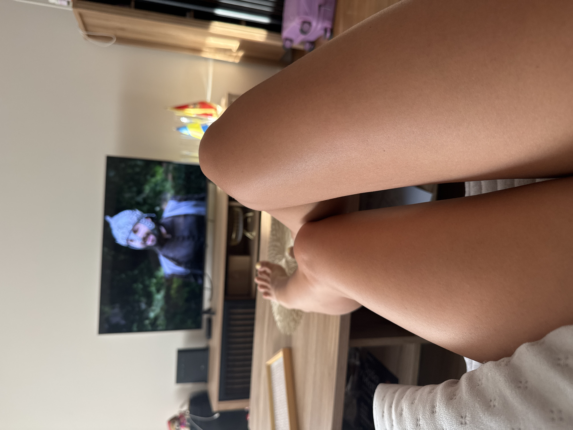 A medium shot captures a person's legs and feet in the foreground, positioned from the knees down. The individual is relaxed, with their legs extended, and their bare feet are visible. In the background, a television screen displays a man wearing a distinctive hat and armor, suggesting a scene from a historical drama or fantasy film. The room appears to be a living area, with a wooden coffee table in front of the TV. On the table, a rectangular tray with a textured insert is visible. To the right of the television, a wooden cabinet stands, and beyond it, sunlight streams through a window, casting warm shadows. A purple suitcase is also visible in the far right of the frame. The overall impression is one of a leisurely moment spent indoors, likely in a domestic setting, with entertainment being the focus.