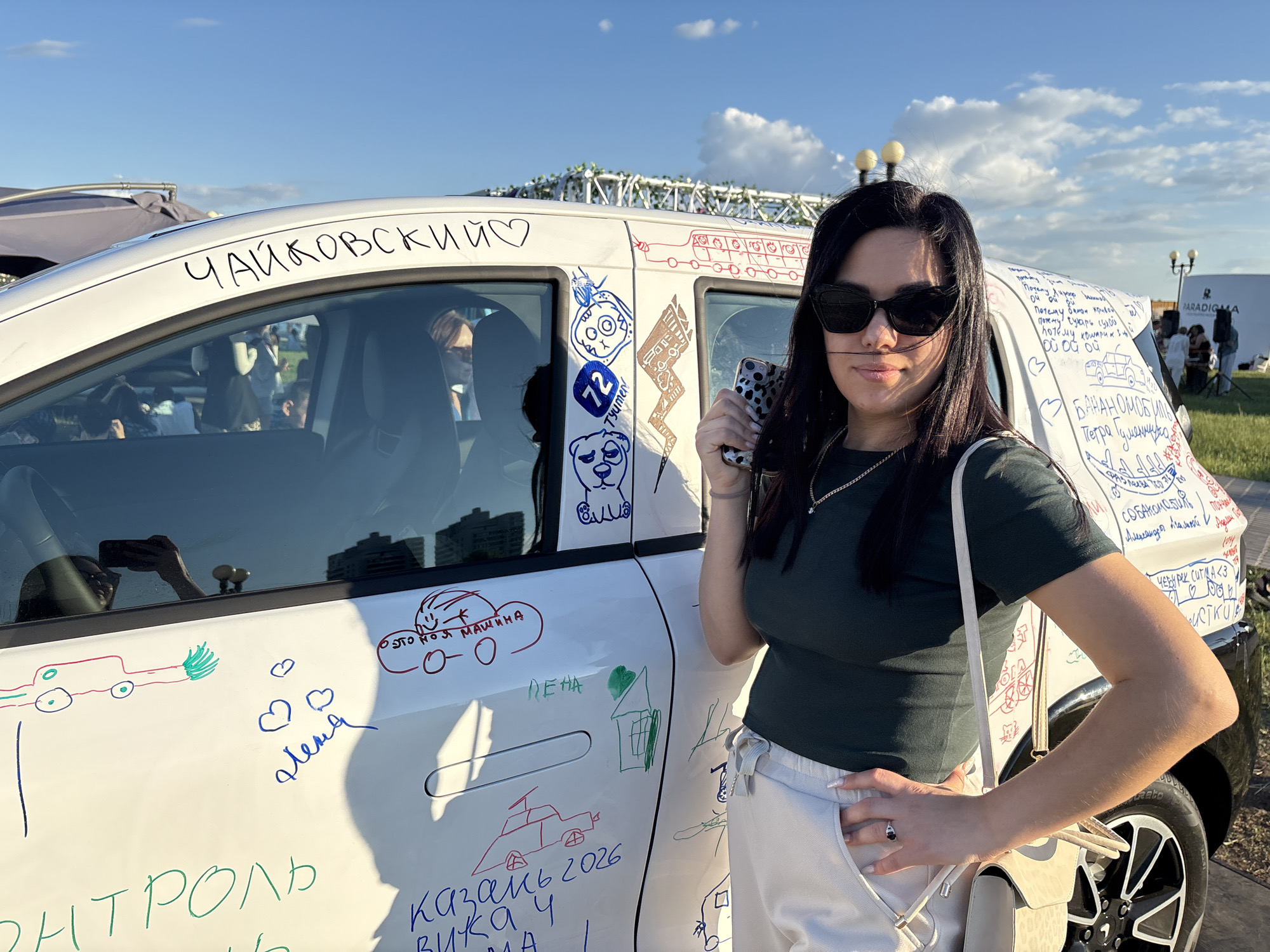 A young woman stands outdoors beside a white car extensively covered in handwritten text and drawings, under a clear blue sky. The woman has dark hair, wears dark sunglasses, a dark green short-sleeved top, light-colored athletic pants, and a light-colored shoulder bag. She holds a smartphone with a patterned case in her right hand, while her left hand is in her pocket, as she faces forward.
The white car, appearing to be a sedan, is marked with various colored markers. Prominent Russian text on the car's roofline reads "ЧАЙКОВСКИЙ" (Tchaikovsky) followed by a heart. On the driver's side front door, "ЭТО МОЯ МАШИНА" (This is my car) and "Алёна" (Alyona) with two hearts are visible. On the rear door, "КАЗАНЬ 2026 ВИКА Ч" (Kazan 2026 Vika Ch) is written in blue, explicitly identifying the location as Kazan, Russia. Other discernible Russian words include "КОНТРОЛЬ" (Control), "БАНАНОМОБИЛЬ" (Bananomobile), "СОБАКОВСКИЙ" (Sobakovsky), and "ТЮМЕНЬ" (Tyumen) alongside the number "72" within a blue circle. The drawings on the car include cartoon faces, a sad dog, stylized vehicles, houses, lightning bolts, and a series of linked circles.
The environment is an open area with green grass under a blue sky dotted with white clouds. In the background, other individuals are visible, along with a white structure bearing the text "PARADIGMA," decorative trellises, and light poles. Tall buildings are reflected in the car's windows.