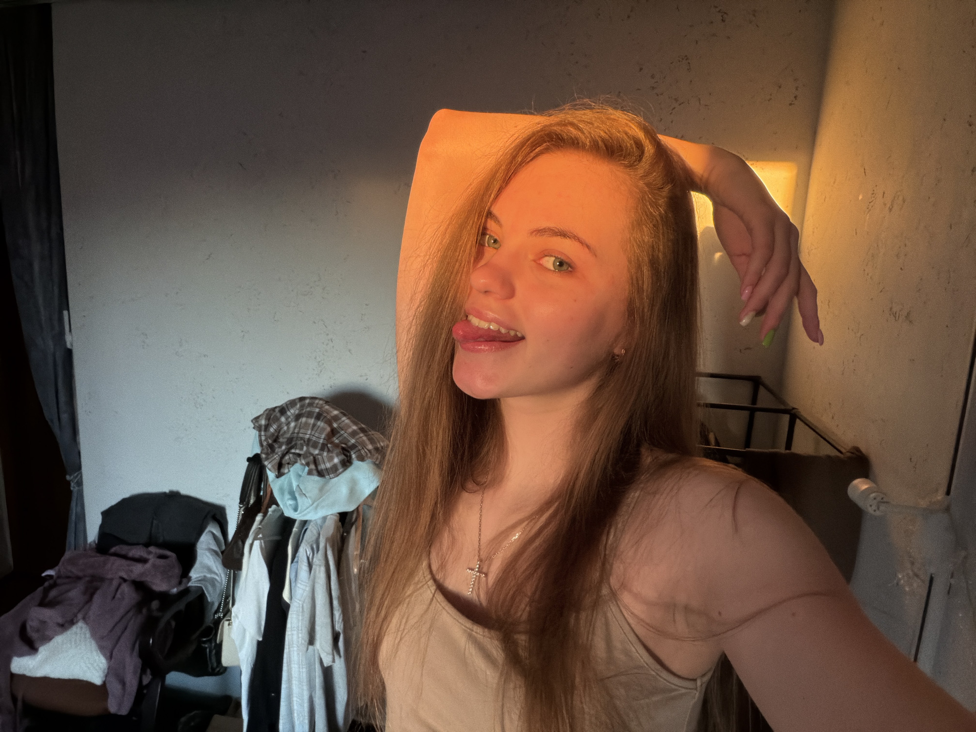 The image is a selfie taken by a young woman with long, light brown hair. She has blue eyes and is sticking her tongue out playfully. She is wearing a light tan tank top and a silver cross necklace. Her right arm is raised and bent behind her head, and her left hand is resting on her thigh. The lighting suggests it is late afternoon or early evening, with warm sunlight casting a glow on her face and arm.
The background appears to be a cluttered room. To the left, there are piles of clothes on what looks like a chair and a rack of clothing hanging on hangers. A dark curtain is visible on the far left. To the right, there is a wall with a textured surface and what appears to be a metal railing or structure.
The overall impression is of a candid, informal moment captured in a personal space. The presence of clothing and the domestic setting indicate the photo was likely taken in a residence. The image does not contain any specific visual cues that would allow for precise localization within the city of Dnipro, Ukraine.