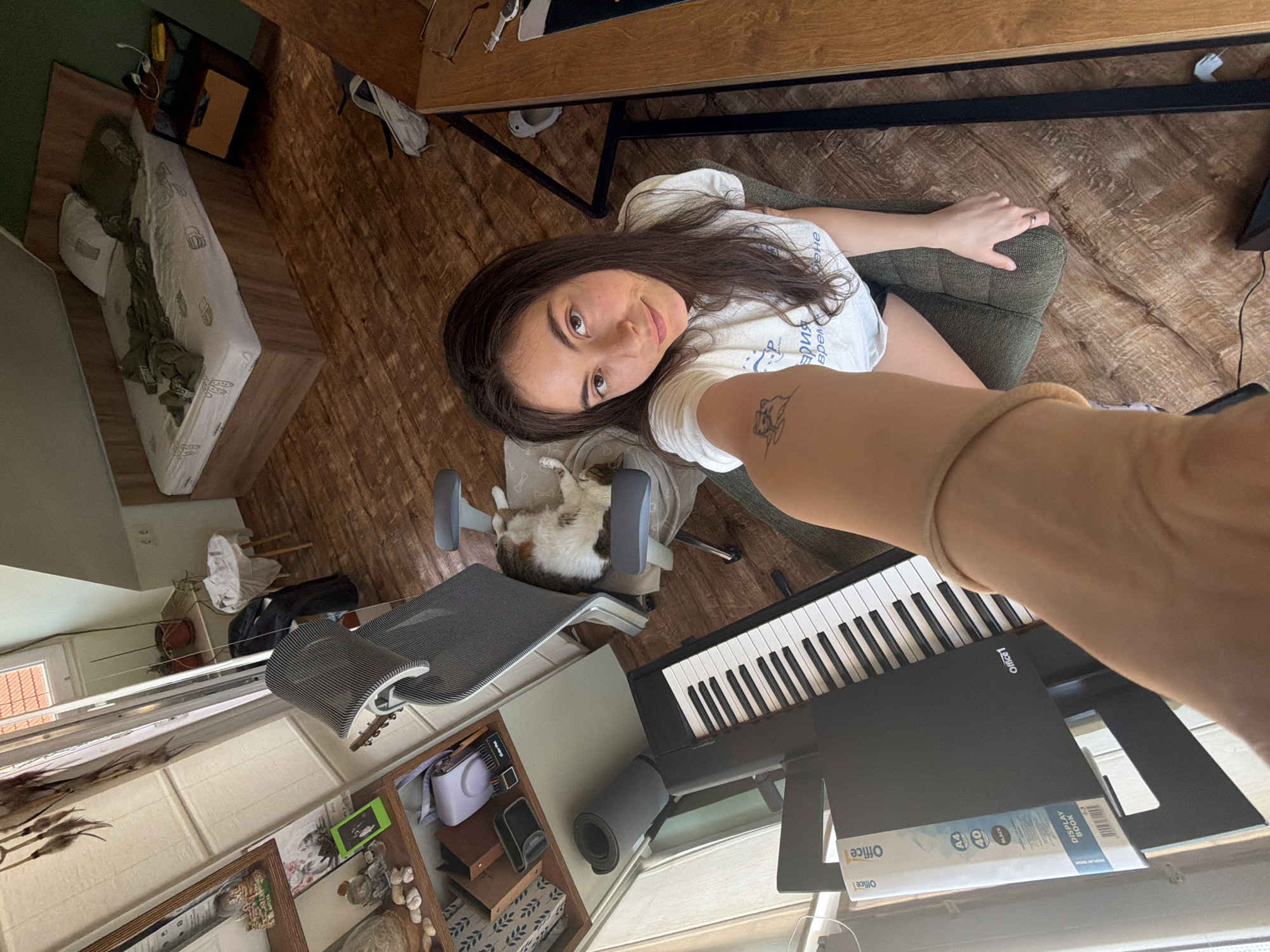 The image captures a young woman taking a selfie in what appears to be an indoor setting. The location is Unknown, Unknown. She is sitting on a green chair near a digital piano. A cat is sleeping on a grey office chair with wheels. The woman is wearing a white t-shirt. The room contains furniture and decor, including a wooden desk and various decorative objects near a window. The image is taken at an angle, providing a partial view of the room's contents.