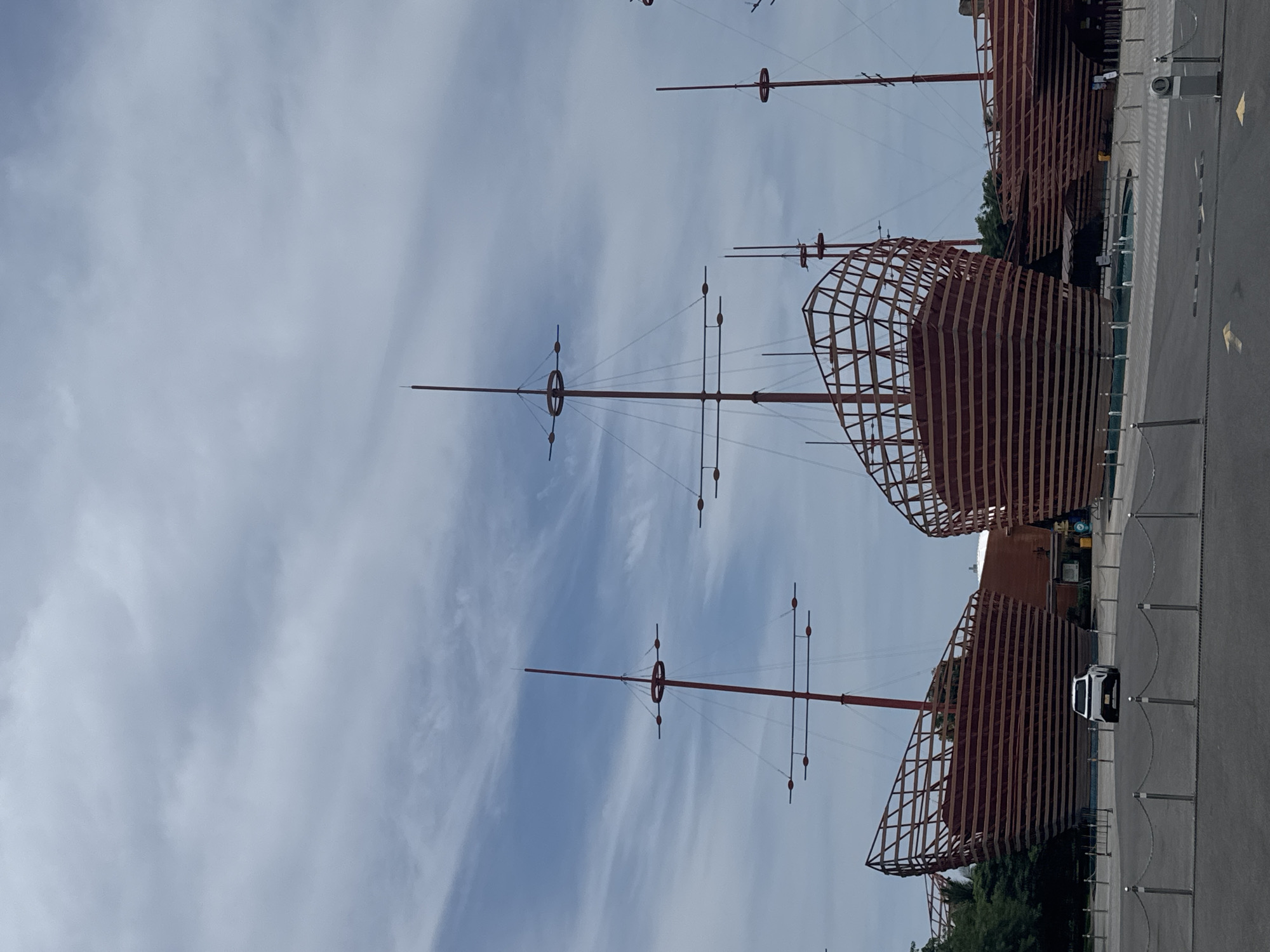 The image shows a nautical-themed structure in Đà Nẵng, Vietnam. The structure comprises skeletal wooden "ships" with tall, slender masts extending into a cloudy sky. The structures have a reddish-brown hue, and their hulls are defined by a lattice of wooden planks. A parking area with a light-colored vehicle is visible next to the structure. The setting is outdoor, with a paved area in the foreground.