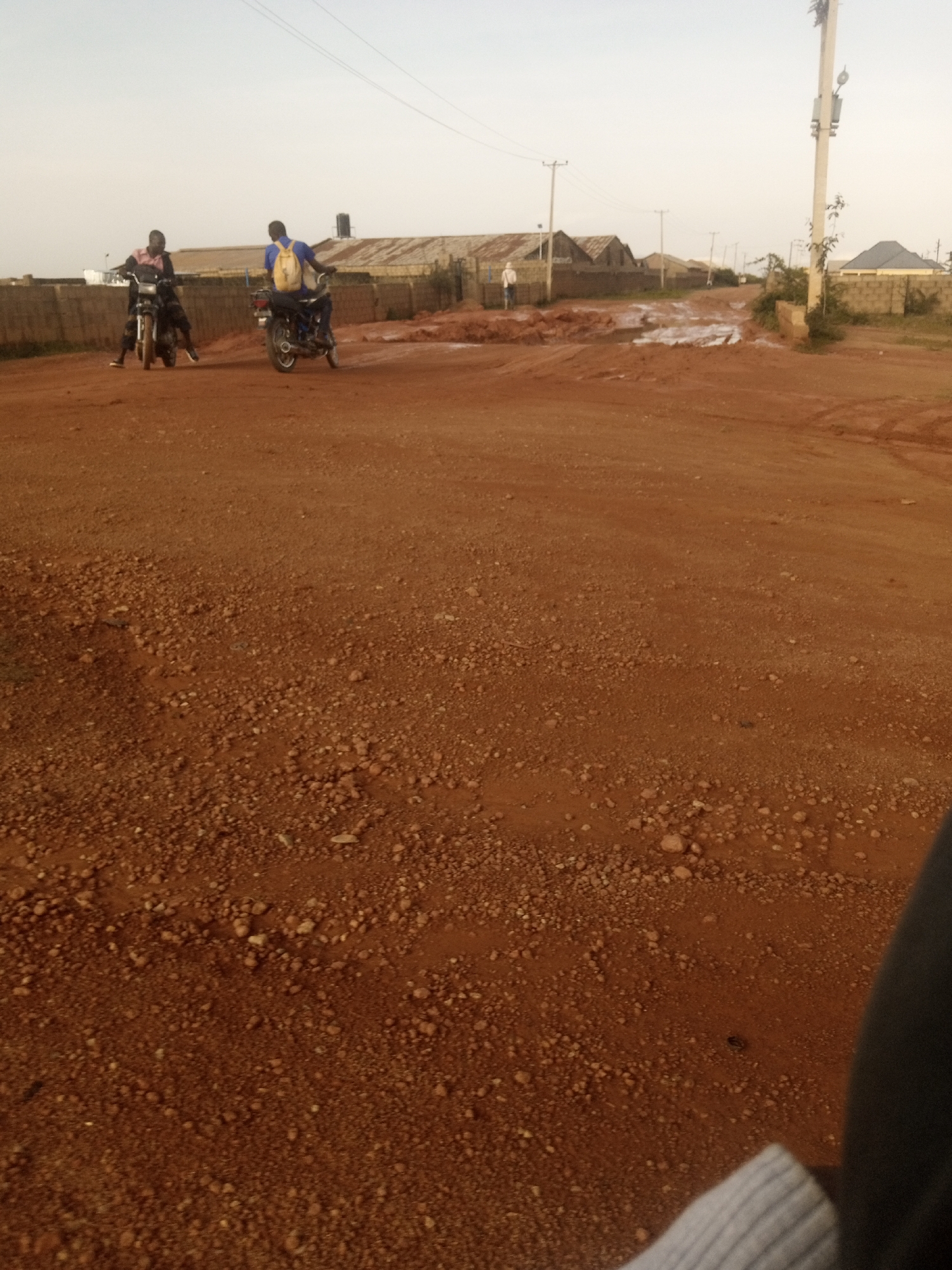 In the city of Jos, Nigeria, two individuals are observed riding motorcycles on a dirt road. The motorcycle in the foreground is ridden by a person wearing a light-colored shirt and dark pants, with a dark-colored motorcycle. Behind them, another individual is on a motorcycle, wearing a blue shirt and yellow backpack, with a dark-colored motorcycle. The road is covered in reddish-brown dirt and gravel, with evidence of puddles and muddy areas, suggesting recent rainfall. In the background, several buildings with corrugated metal roofs are visible, along with a utility pole and electrical wires. The sky is overcast with a pale hue.