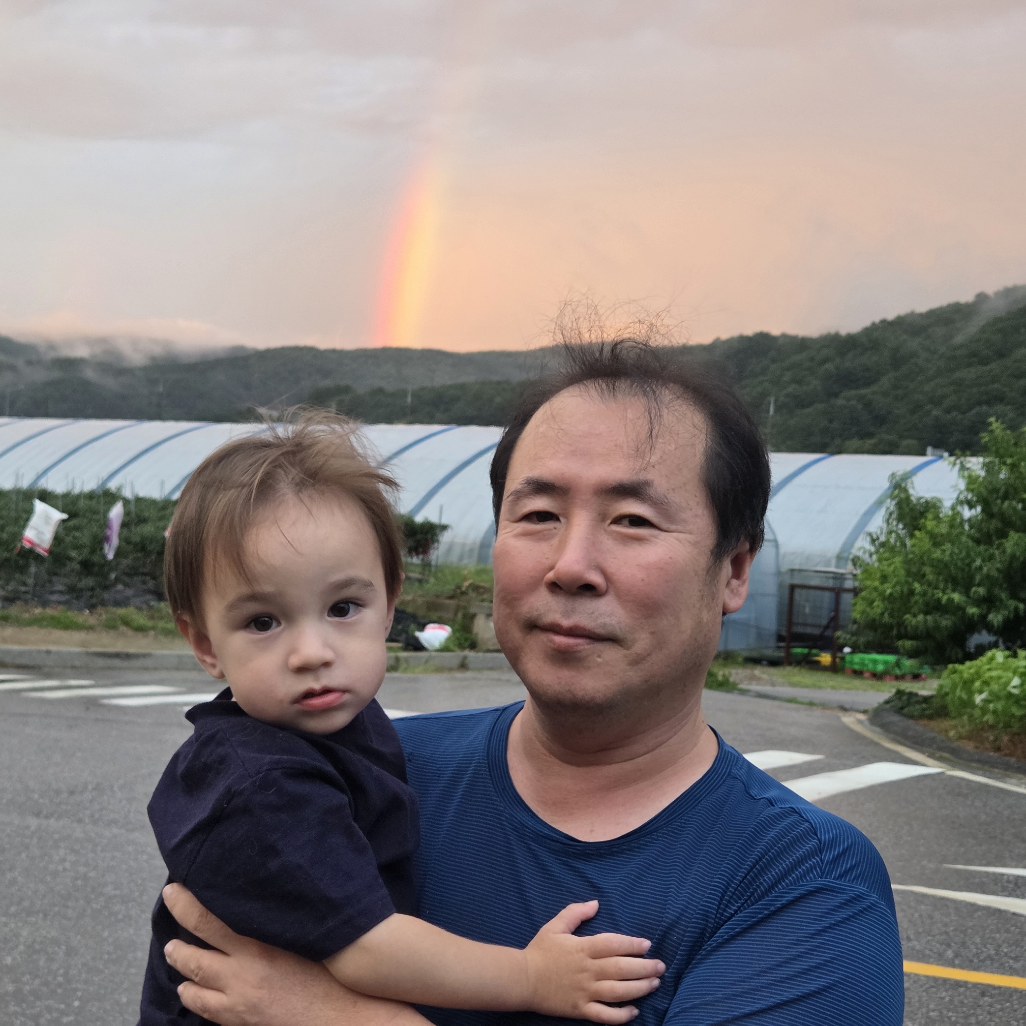 A man in a blue striped t-shirt holds a young child in front of a rainbow in Dunnae-myeon, South Korea. The child, with light brown hair, is wearing a dark blue shirt. The man has dark hair and is looking towards the camera. In the background, there are green hills covered in trees, a large white greenhouse-like structure, and a road with white crosswalk markings. The sky is cloudy with a faint pink and orange hue, and the rainbow is visible in the upper left portion of the frame.