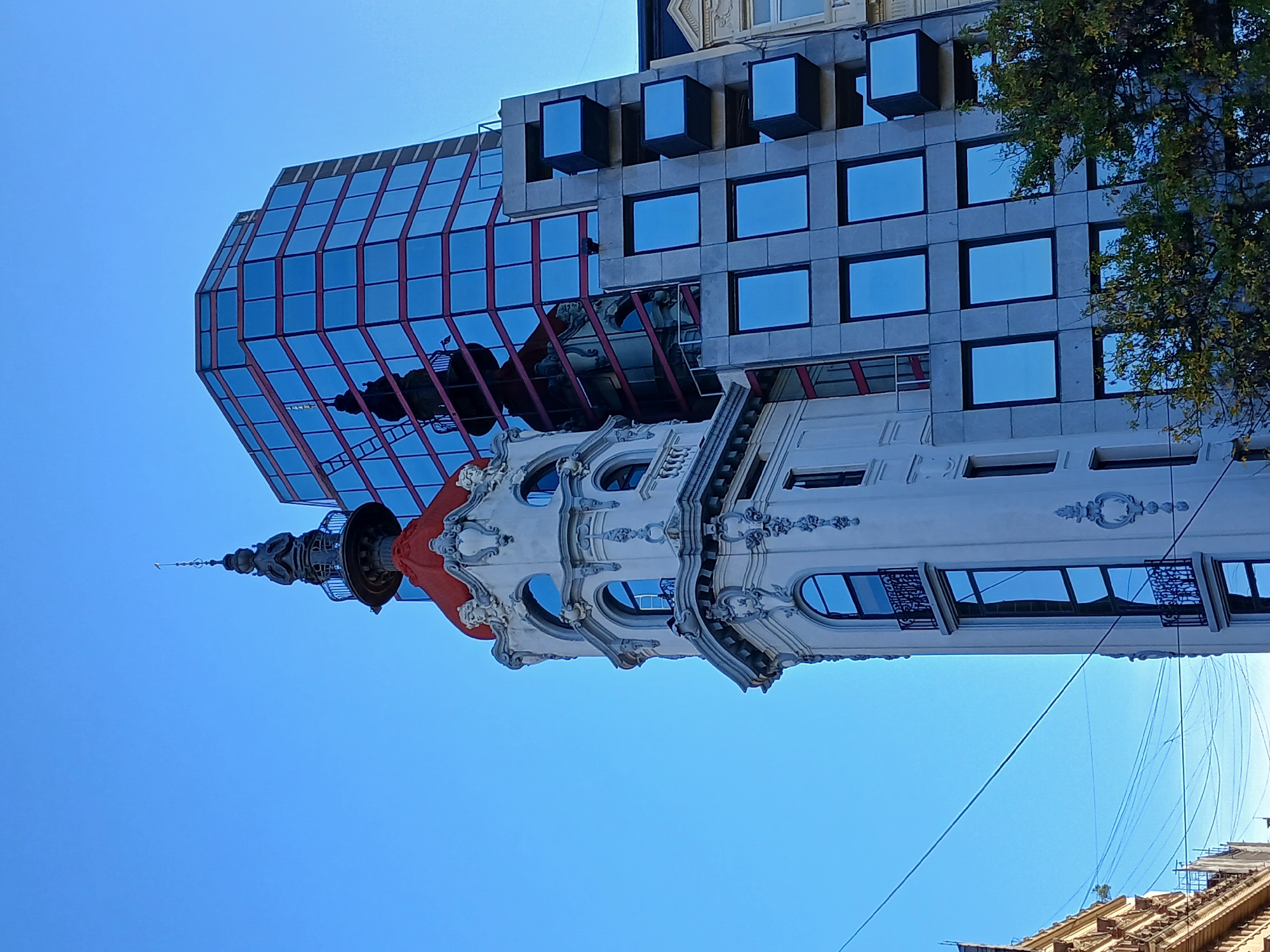The image depicts a building facade in Buenos Aires, Argentina. It is a combination of architectural styles, with a highly ornamented, white, historical building at its base. The historical building features arched windows, decorative moldings, and a red, bulbous rooftop structure. Above the historical building is a modern structure with a glass facade and red framing elements. The modern building also has sections with square windows that appear to be projecting slightly outward. The sky is clear and blue. Power lines are visible in the bottom right corner of the image.