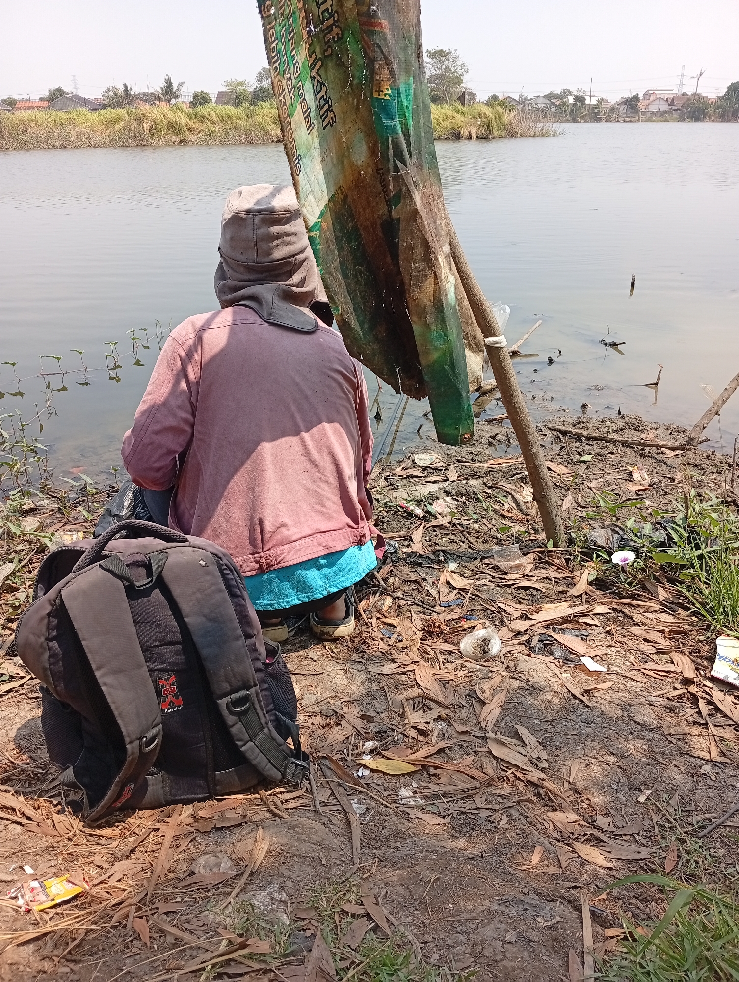 A person wearing a light-colored hat and a pink shirt with a blue waistband sits facing a body of water. A black backpack rests on the ground in front of them. The scene appears to be an outdoor, possibly rural, location in Karangmukti, Indonesia, near a body of water. The ground is covered in dry leaves and debris. There is a makeshift shelter made of fabric and sticks behind the person. The water's edge is muddy, with some small plants growing near it. The sky is clear and bright, suggesting it is daytime.