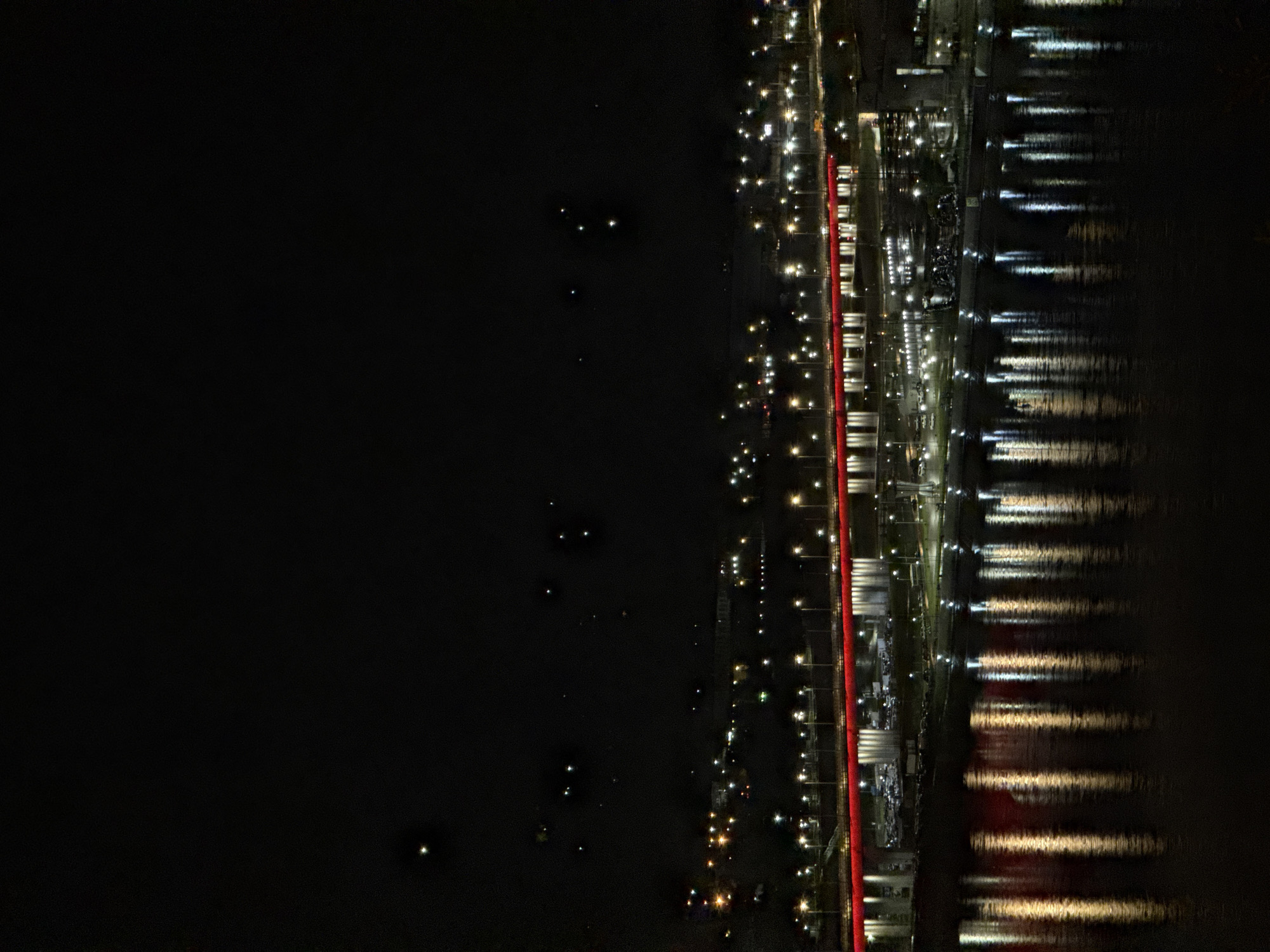 The image shows a nighttime view of a city on the edge of a body of water, likely a river. The city's edge is delineated by a row of lights, a distinct red line, and various buildings reflecting light. The water surface shows multiple light reflections. The darker, left side of the image suggests an undeveloped area or simply the unlit portion of the river. Location of Krasnoyarsk, Russia is inferred by the bridge known to be the Vinogradovsky Bridge.
