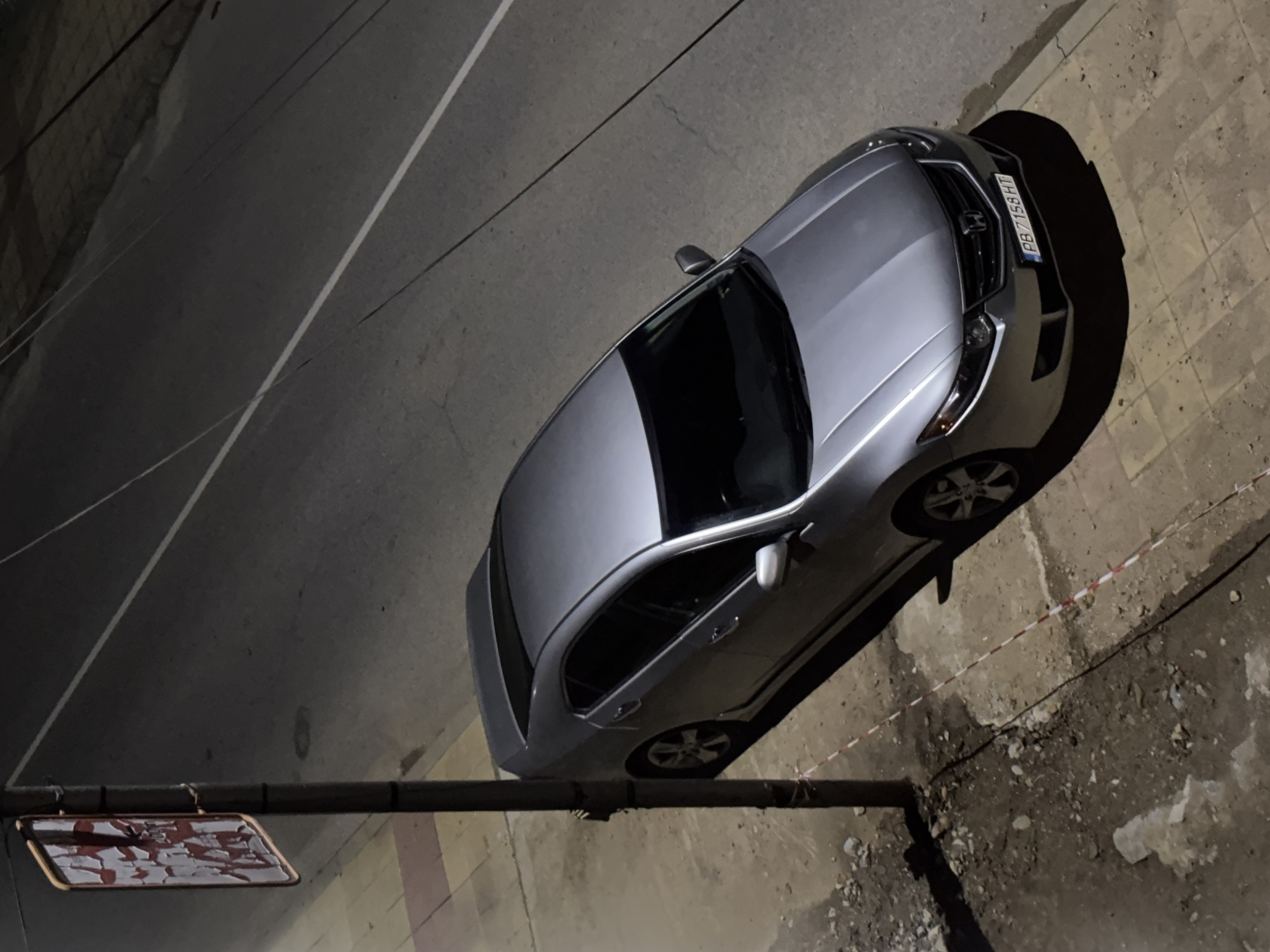 The image is an aerial shot of a silver car parked on a street. The car is positioned on the right side of the frame, occupying a portion of the road and sidewalk. The street surface appears to be asphalt, with white painted lines marking the lanes. A street sign is visible on the lower left corner of the image. The sidewalk is uneven with patches of dirt and debris. The license plate number of the car is PB 7158 HT. The scene is dimly lit, suggesting it is taken at night. The location is Zlatograd, Bulgaria.