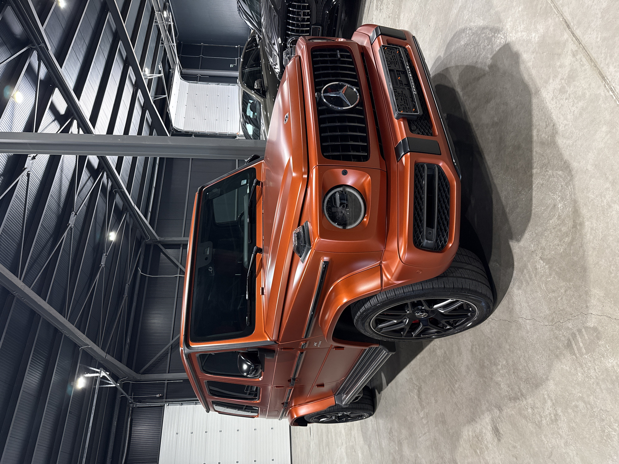 An orange Mercedes-Benz G-Class is parked in a clean, modern industrial-style garage, possibly a showroom or storage facility, with a grey concrete floor and a dark metal ceiling with exposed beams and fluorescent lighting. The garage features large white roll-up doors in the background. Two other dark-colored vehicles are partially visible behind the G-Class. The G-Class has black accents, including the grille, wheels, and side mirrors, and a license plate frame is attached to the front bumper. The image is a medium shot, focusing on the front-right quarter of the vehicle. There are no people visible in the image. The garage is well-lit and appears to be in an urban or industrial area. This scene is not specifically located in Plovdiv, Bulgaria, and no information in the image suggests this location.