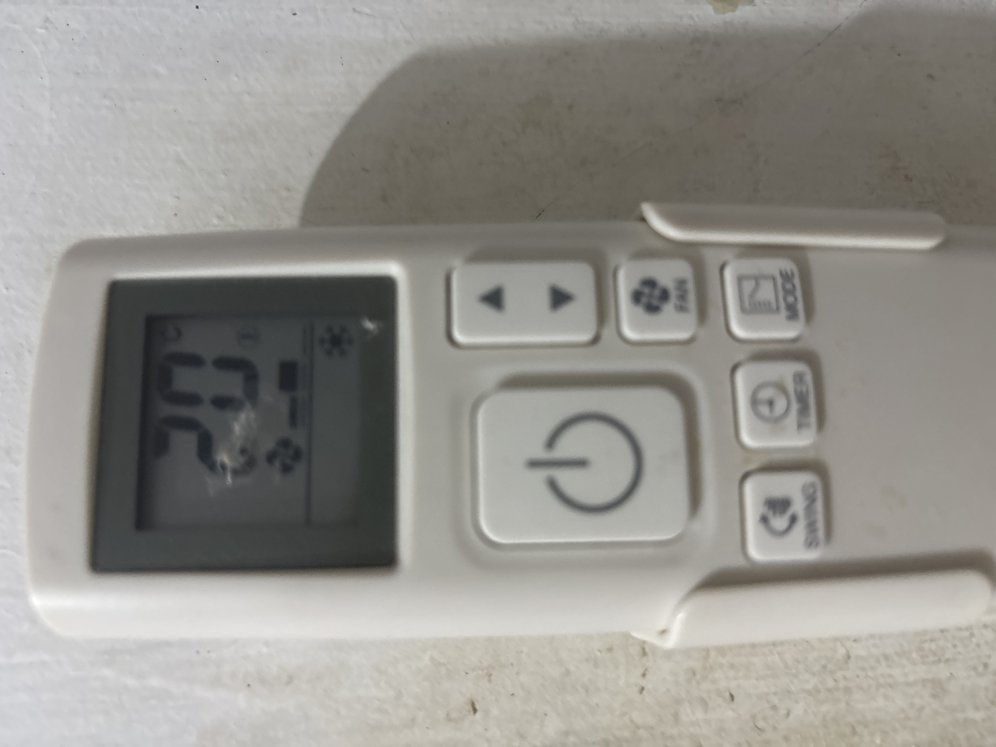 A white remote control for an air conditioner is held by an unseen person against a textured white wall. The remote has a digital display showing "20°C" and various icons, including a power button, up and down arrows, a fan, swing, timer, and mode buttons. The remote is positioned vertically, and the lighting suggests an indoor environment. The wall has some faint marks and shadows. There is no indication of the city of Geulanggang Teungoh, Indonesia within the image itself.
