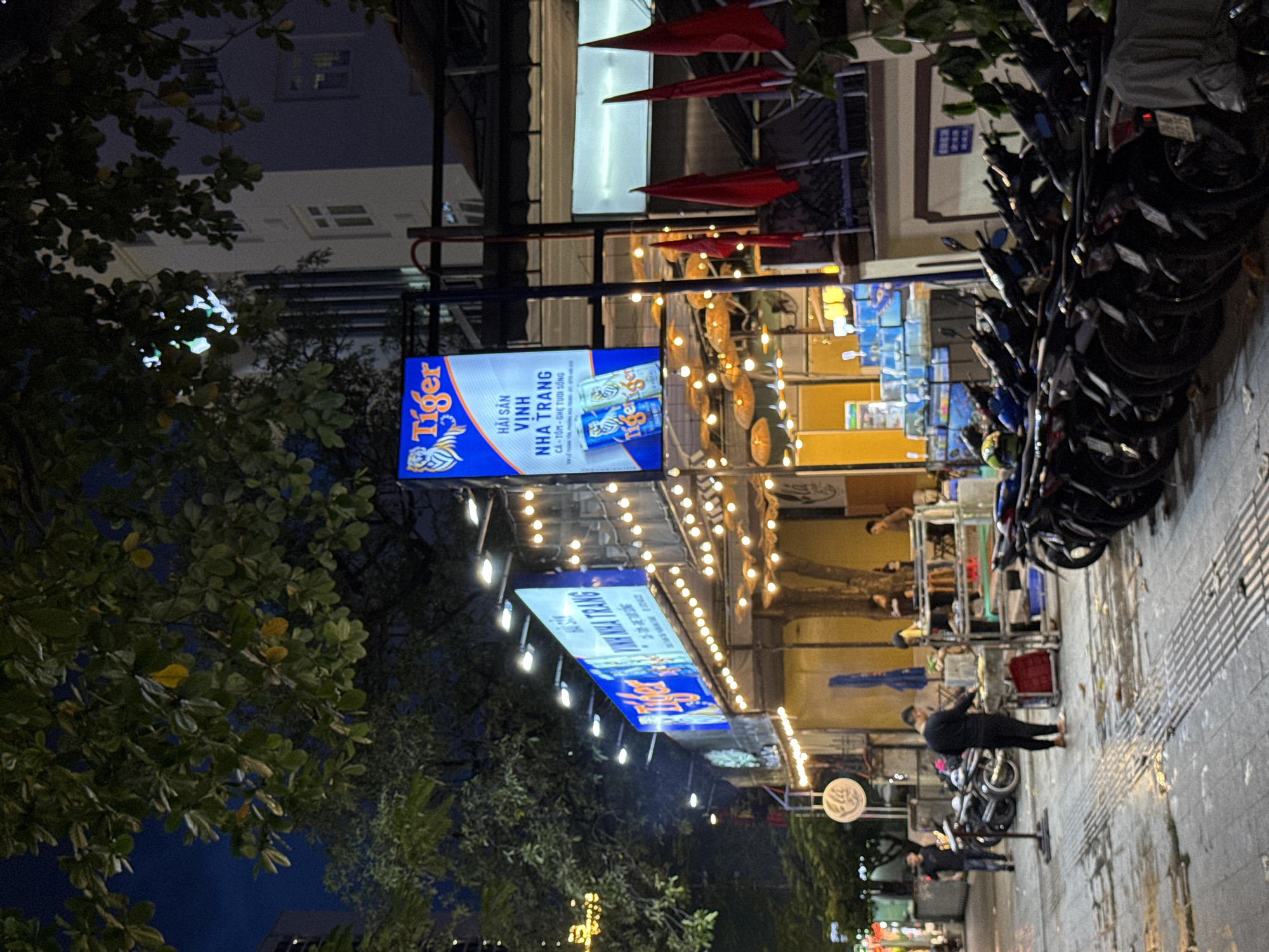 The image depicts a street scene at night in Nha Trang, Vietnam, likely near a seafood restaurant. In the foreground, a sidewalk is visible, wet with what appears to be recent rain. Several motorbikes are parked along the side of the street. A man in dark clothing is standing near a food cart, preparing food. In the background, a restaurant with bright lights and signage is prominent. A large blue sign with the Tiger beer logo and text in Vietnamese reading "HẢI SẢN VINH NHA TRANG" indicates the establishment is a seafood restaurant in Nha Trang. Red flags are also visible, likely part of a display or decoration for the establishment. The overall atmosphere is lively and suggests a popular dining and gathering spot.