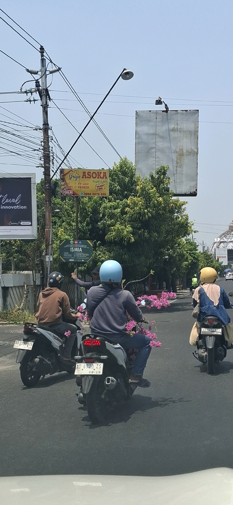 On a bright, sunny day in Surakarta, Indonesia, three motorcycles are seen traveling on a road. The first motorcycle, on the left, is ridden by a person wearing a brown hoodie and a black helmet, with a pink flower attached to the front of the vehicle. The second motorcycle, in the center, is ridden by a person wearing a grey long-sleeved shirt, blue jeans, and a light blue helmet, carrying a large amount of pink flowers. This rider is gesturing with their left hand. The third motorcycle, on the right, is ridden by a person wearing a blue outer garment and a yellow helmet, also carrying pink flowers. In the background, there are utility poles with numerous wires, a street lamp, and various signs, including one for "GRIYA ASOSKA" and another for "ISMIA". Trees and buildings are also visible, suggesting an urban or semi-urban environment. The sky is clear and blue.