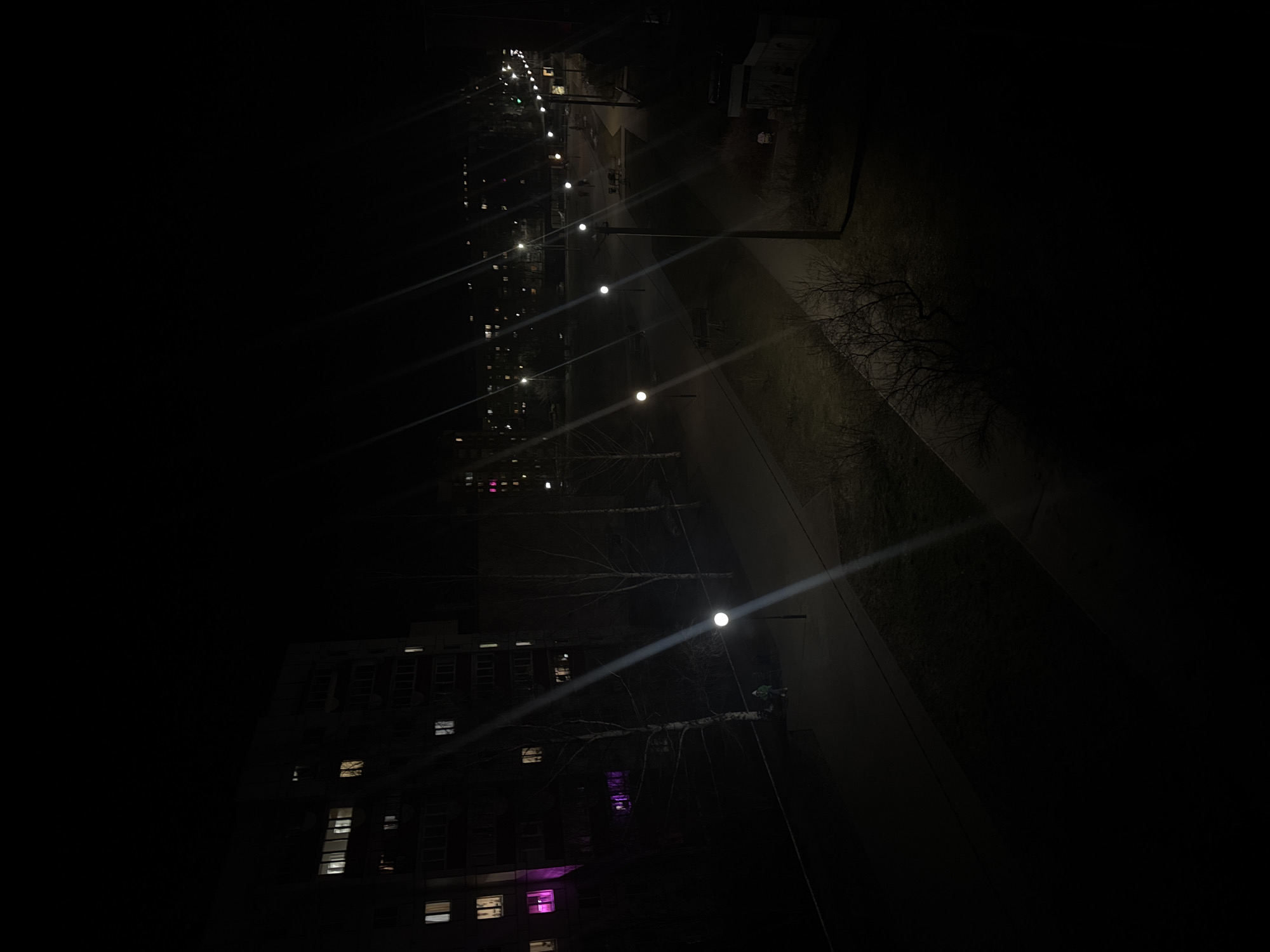 The image depicts a nighttime urban scene, possibly in Chaikovsky, Russia, characterized by a stark contrast between illuminated areas and darkness. A multi-story building with several lit windows, including some with a distinctive purple hue, is visible. A row of streetlights illuminates a pathway or road, casting bright beams of light through the darkness. Bare trees are silhouetted against the limited light. The overall atmosphere is quiet and still.