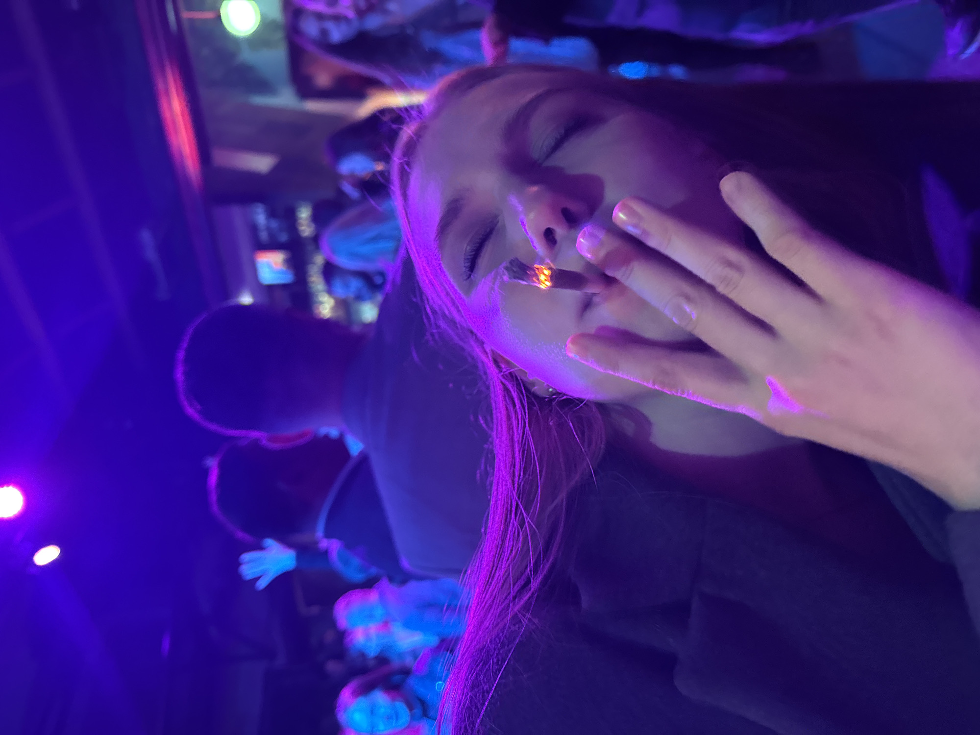 The image shows a close-up of a young woman with long, auburn hair, illuminated by purple and pink lights in what appears to be a dimly lit club or bar. She is holding a lit cigarette to her mouth, with her eyes closed and a slight smile on her face. Her right hand is raised, partially obscuring her face, and her fingernails are painted with a light, shimmery polish. In the background, other people are visible, some with their hands raised, suggesting a lively atmosphere, possibly a concert or a party. The lighting and attire of the people in the background suggest a social gathering. There is no specific indicator that this location is in Perm, Russia.