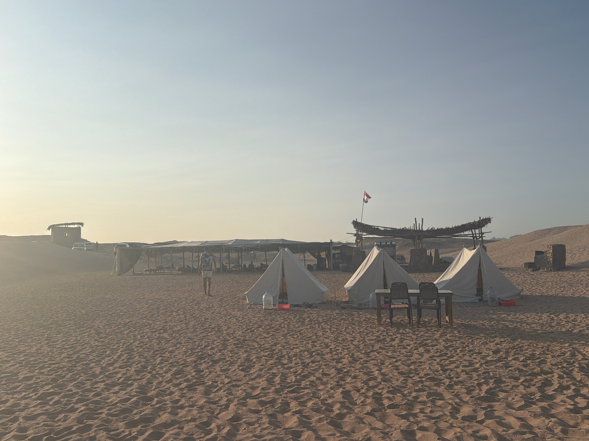 The image depicts a desert landscape with a campsite in Sharm El Sheikh, Egypt. The foreground is dominated by rippled sand dunes. In the mid-ground, three white canvas tents are set up, with a table and chairs placed between two of them. A person wearing a backpack and shorts walks away from the viewer towards the tents. In the background, there is a shaded area with tables and chairs, and a small structure on a dune. Several cars are parked near the structure. A flagpole with a flag is visible on a raised structure in the distance, and a rocky outcrop is also present. The sky is clear and light, suggesting either early morning or late afternoon.