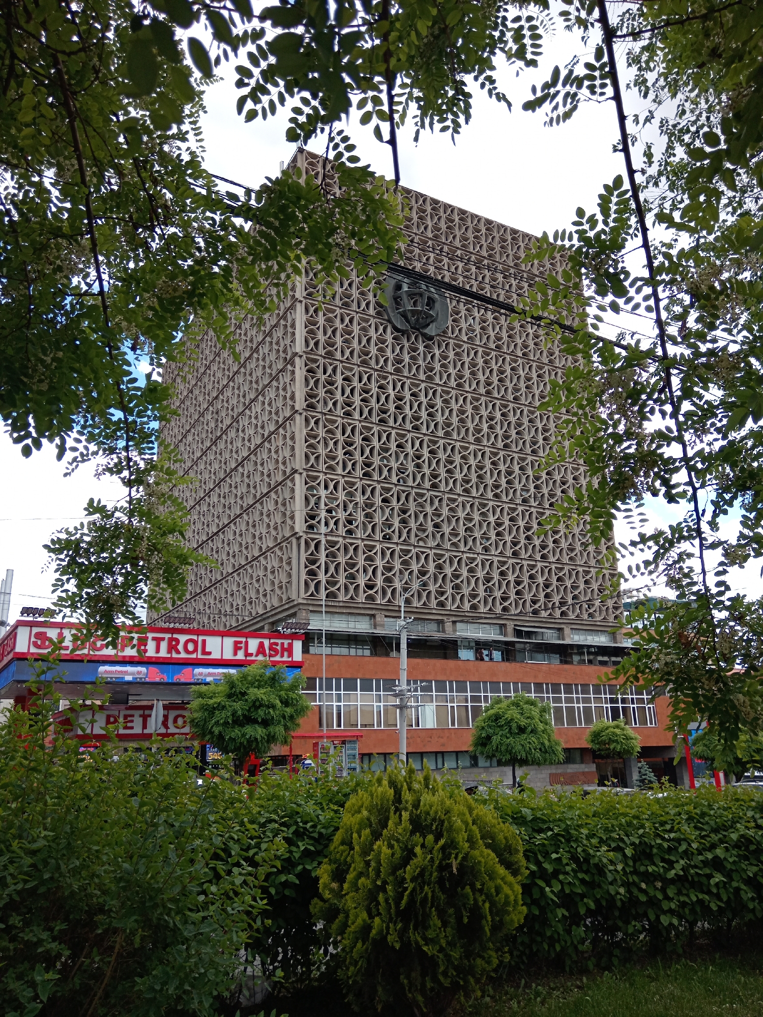 A tall building with a geometric facade is visible through tree foliage and above a gas station. The gas station is labeled "PETROL FLASH." The building appears to have a crest near the top and is situated above a lower, red brick building with many windows. Green bushes and lawn are in the foreground. The location is in the city of Unknown, Unknown.