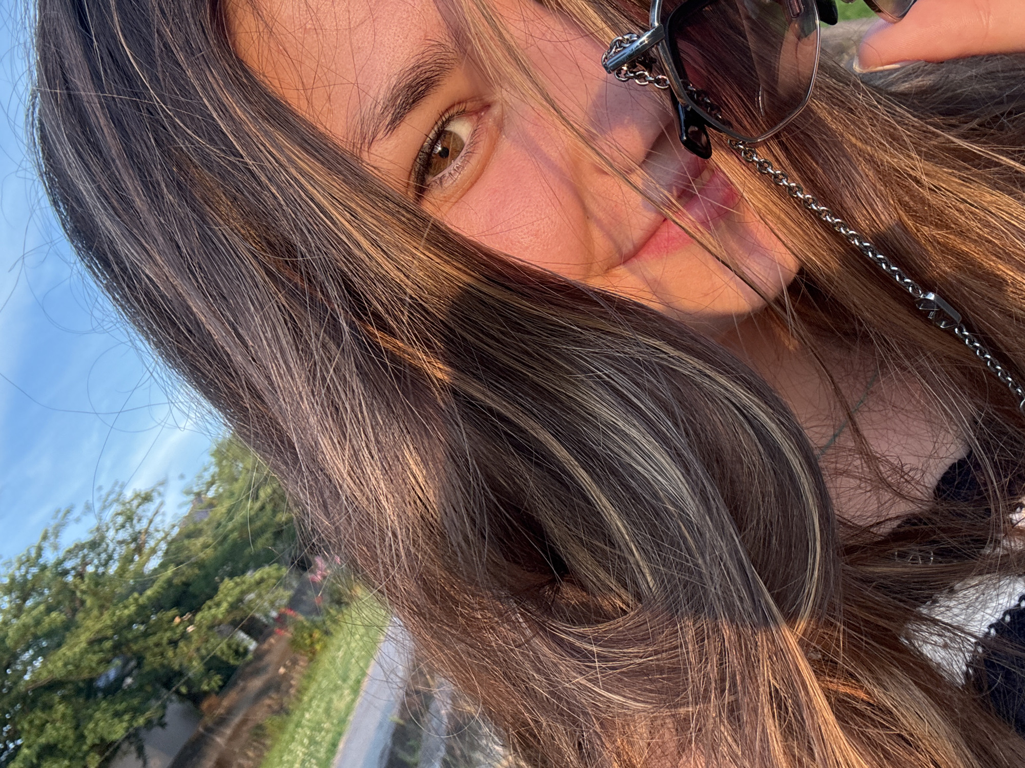 A close-up, eye-level outdoor selfie depicts a young woman smiling in golden hour sunlight in Kanazawa, Japan. Her face is partially obscured by her long, brown hair with blonde and caramel highlights, which also casts soft shadows. Visible features include her right eye, which is brown, part of her nose, and her pink-lipped mouth curved in a smile. Her right cheek exhibits a warm, rosy flush from the direct sunlight. She is wearing dark-lensed sunglasses with a black frame, which are suspended by a prominent metallic chain around her neck; the chain features a distinct 'V'-shaped emblem. A finger, presumably her right index finger, is visible near the bottom right, lightly touching the sunglass frame. The background, blurred, reveals a clear blue sky with some wispy clouds at the top left, and green foliage, including trees and grass, suggestive of a park or natural outdoor setting, with a path or road also visible to the left.