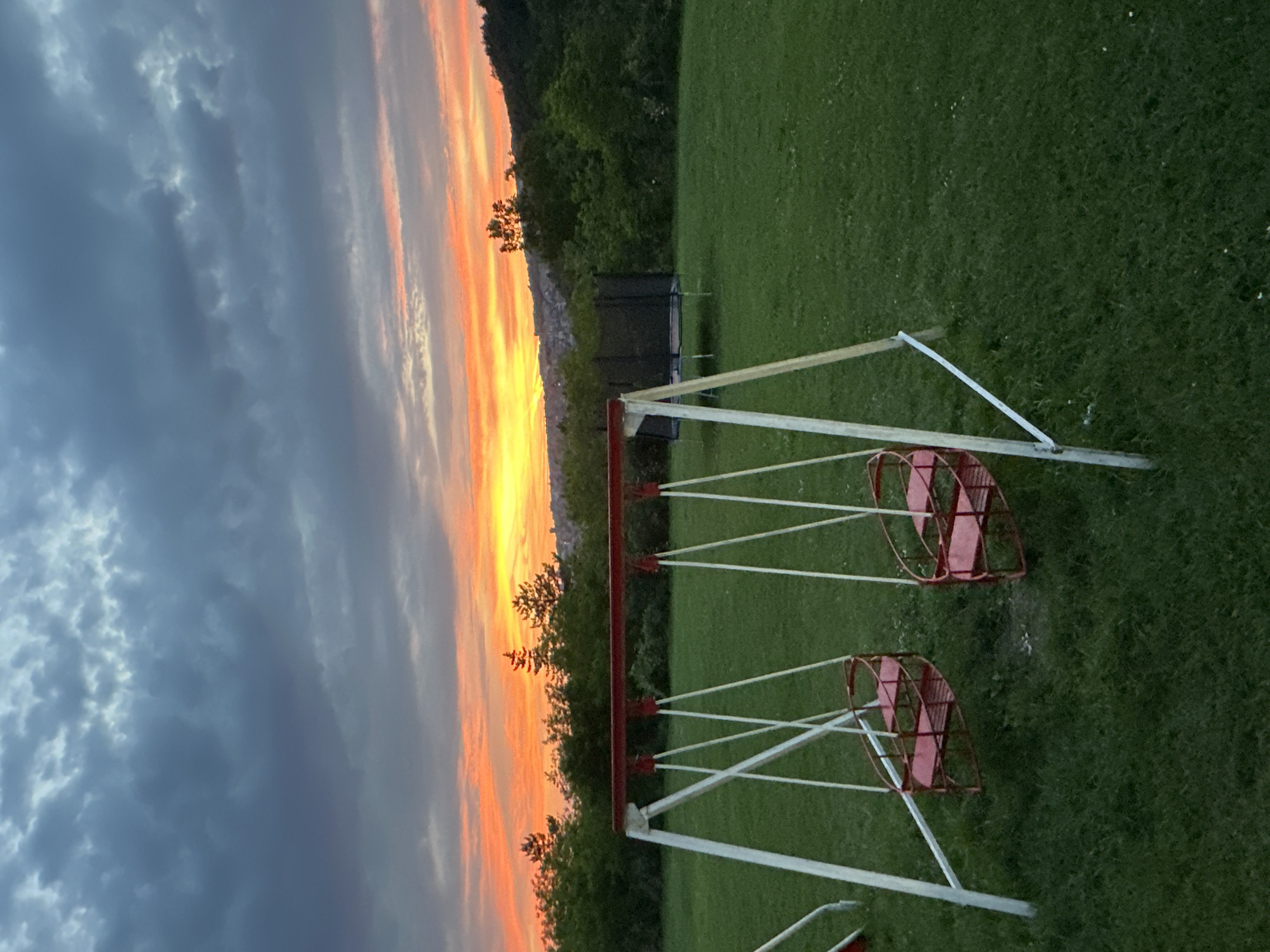 The image depicts a playground with a red and white swing set in the foreground, set against a backdrop of green grass and trees. Beyond the trees, an orange and yellow sunset stretches across the sky, meeting dark, grey clouds on the left side of the frame. A trampoline is visible in the background among the trees. The location is likely a park or recreation area near Veliko Tarnovo, Bulgaria, though specific city details are not visually evident.