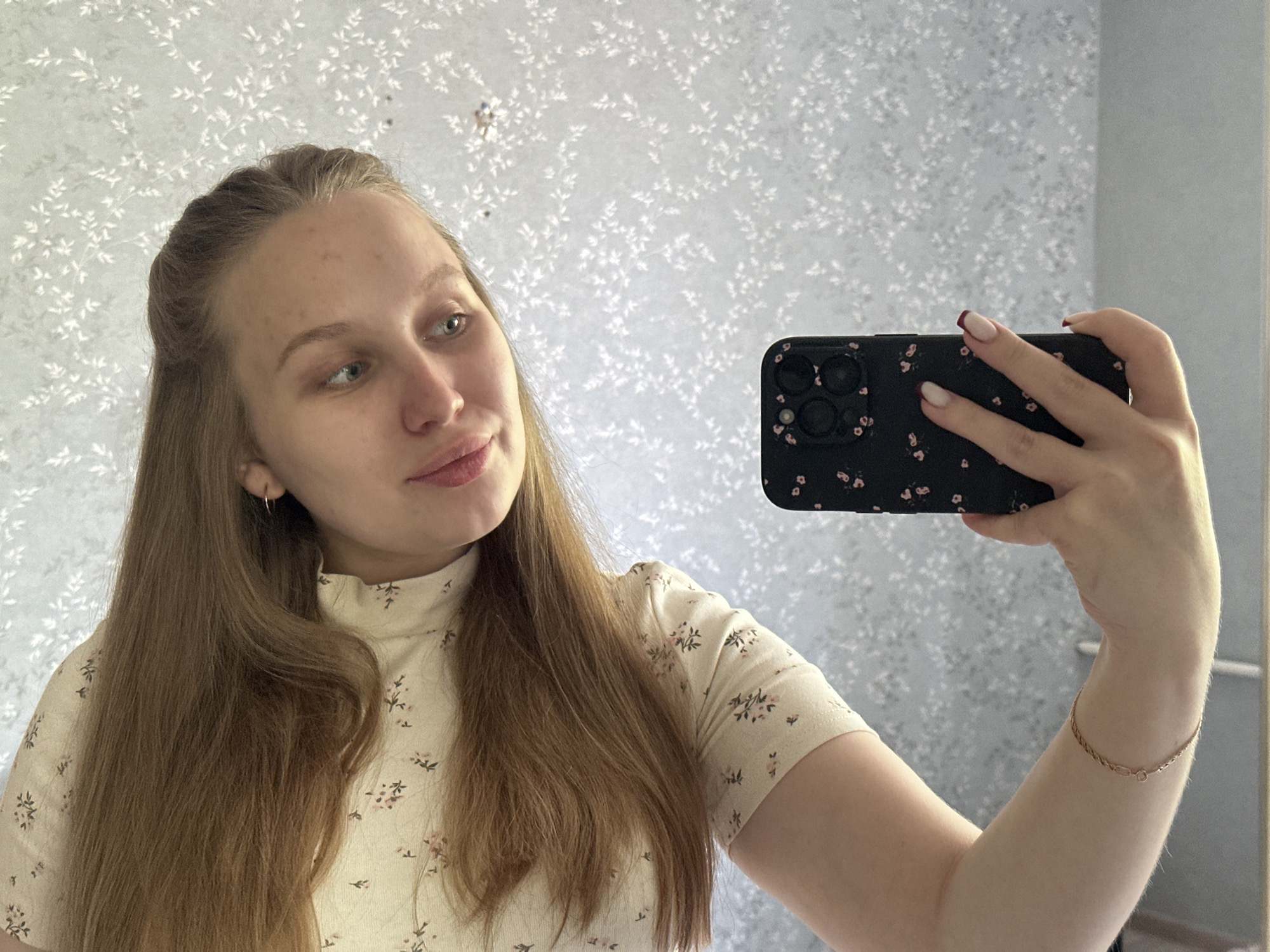 A young woman takes a selfie in front of a patterned wall. She is wearing a light yellow floral top. She holds a black smartphone with a floral case. She has long brown hair, parted in the middle and styled back from her face. She is wearing a gold bracelet. The location is an interior with wallpaper. The city is Chaikovsky, Russia.