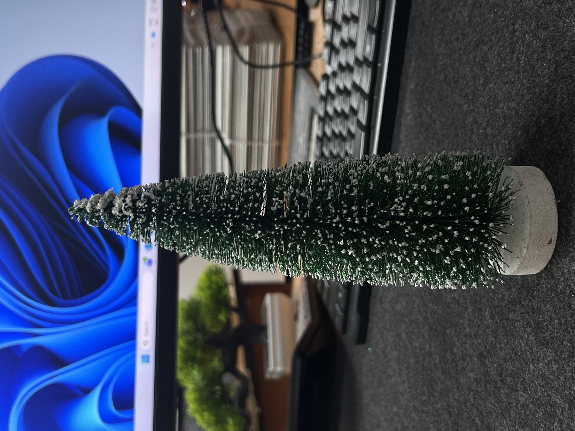 The image shows a close-up, eye-level, indoor shot of a miniature artificial Christmas tree with white tips, placed on a dark gray, textured surface. In the background, a computer monitor displays a vibrant blue abstract wallpaper. To the left of the tree, a small artificial bonsai tree in a white pot is visible. Further in the background, stacks of papers and a black keyboard can be seen. The lighting is dim, suggesting an indoor environment. The image does not depict any people, actions, or interactions. The location is not identifiable as Kadamjay, Kyrgyzstan, as there are no distinct geographical or cultural markers present.