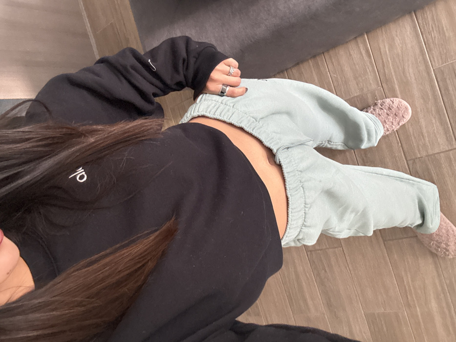 This image, taken in an indoor residential or private setting within Mexico City, Mexico, presents a high-angle, downward view of an individual from the mid-chest to the feet. The subject, likely female, wears a black cropped long-sleeve sweatshirt with 'alo' in white script on the left chest and a small white hook-like emblem on the left cuff. Below the sweatshirt, a section of the abdomen, including the navel, is visible. Light mint-green elastic-waist sweatpants are worn on the lower body. The subject's feet are clad in light pink, textured, closed-toe slippers. Long, straight dark brown hair falls over both shoulders, partially obscuring the upper body. A portion of the lower face, exhibiting red lipstick, is visible at the top edge. The right hand, partially visible, displays a wide, dark ring on the index finger and a thin, potentially jeweled, ring on the ring finger. The floor consists of light brown wood-look tiles, with a dark gray rug or mat partially visible on the right side of the frame.