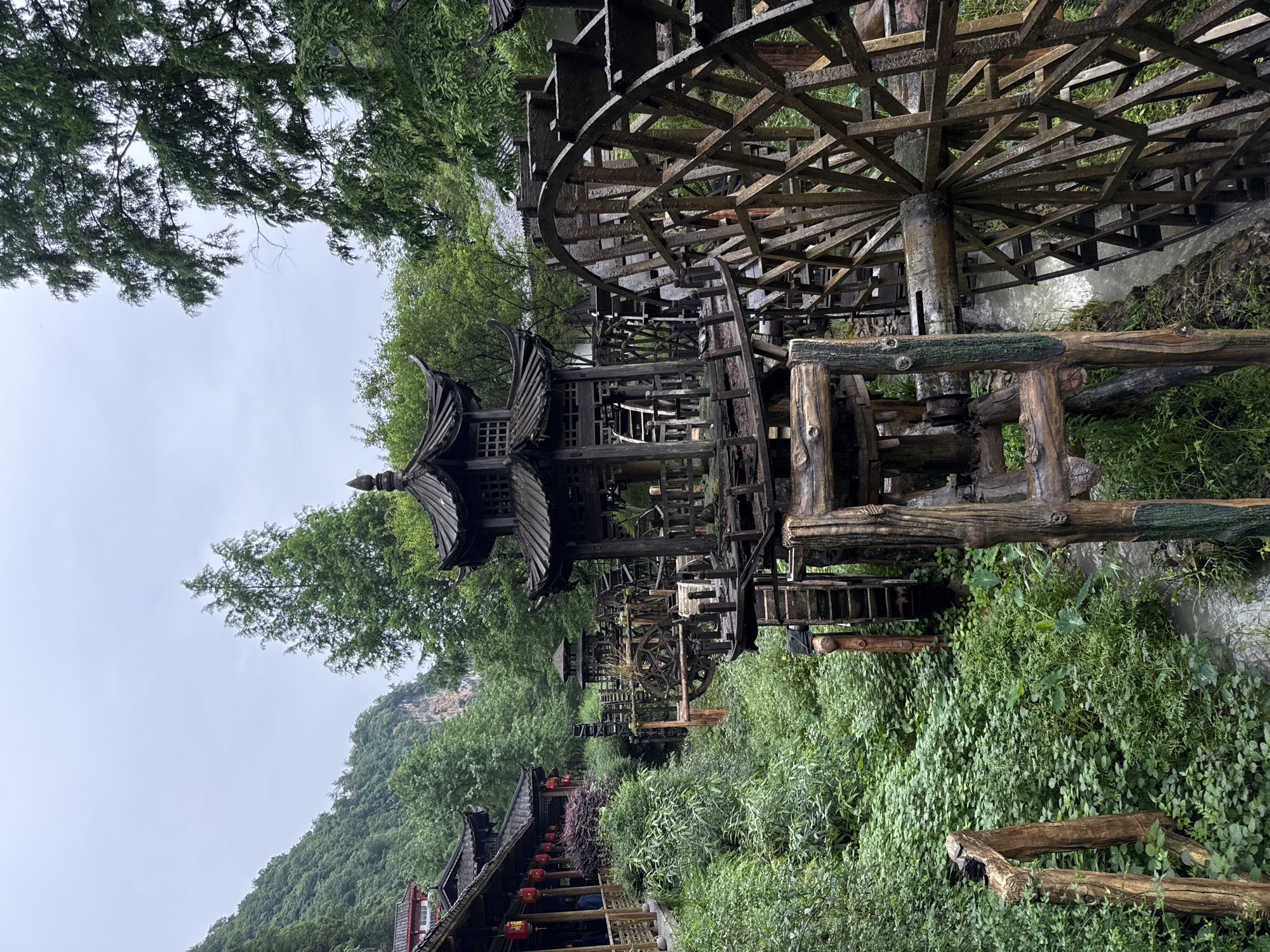 The image depicts a scene in Jundiping, China, featuring a series of traditional wooden water wheels along a stream. The water wheels are constructed from dark wood and exhibit a lattice-like structure. Interspersed between the water wheels are small pagoda-style structures with tiered roofs. The surrounding environment is lush with greenery, including dense foliage and trees covering a mountain in the background. In the lower left, a building with a traditional Chinese roof and red lanterns is visible. The sky is overcast.