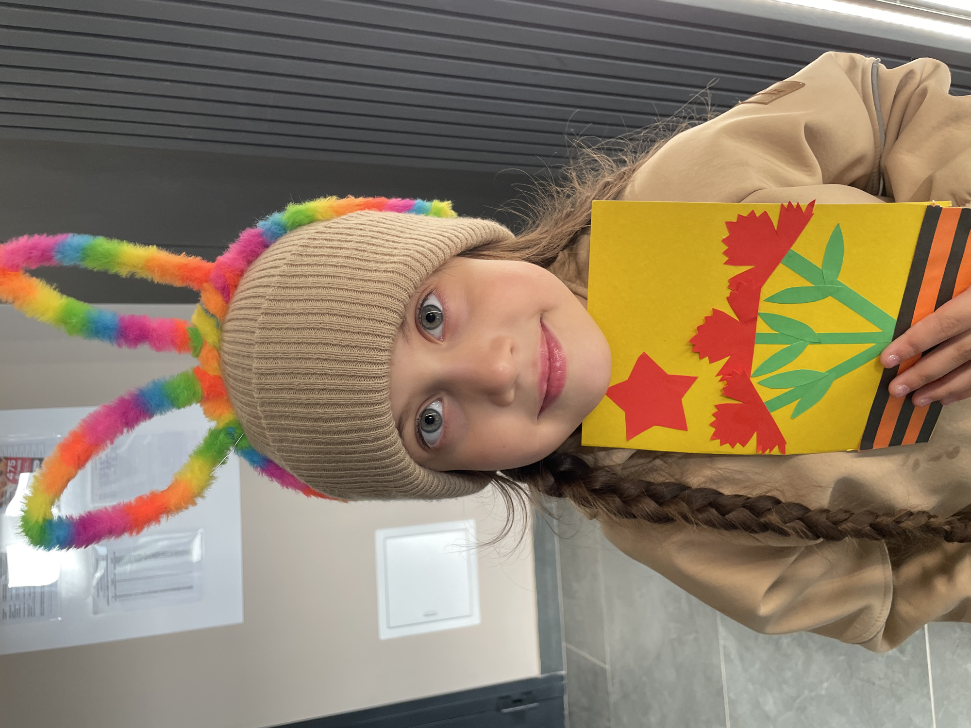 A young girl with light-colored hair braided into a plait over her shoulder is holding a yellow card decorated with red paper flowers, a red star, and a ribbon with black and orange stripes. She is wearing a beige knitted hat adorned with rainbow-colored pipe cleaner loops. The background consists of an indoor setting with a light-colored wall, a gray tiled wall, and a black ventilation unit along the lower left of the frame. The location is possibly in Izhevsk, Russia, based on the decorative elements on the card.