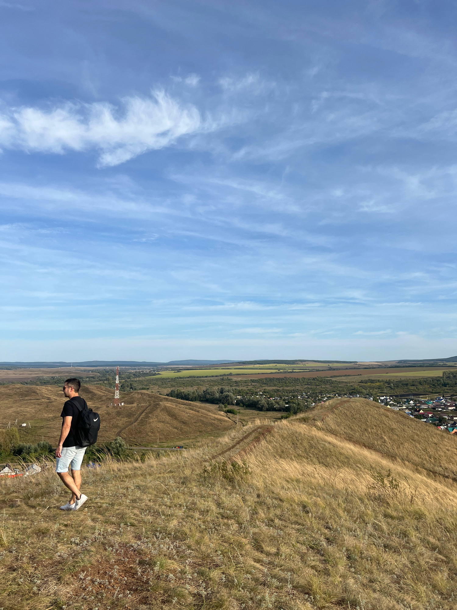 A young man wearing a black t-shirt, light-colored shorts, and white sneakers stands on a grassy hillside overlooking a valley. He has a black backpack on. In the middle ground, there is a radio tower, and beyond that, a town with houses and fields. The sky is blue with scattered white clouds. The man is facing left, looking out at the landscape. The hillside is covered in dry, golden grass, and the ground is uneven. The overall scene suggests a rural or semi-rural setting.