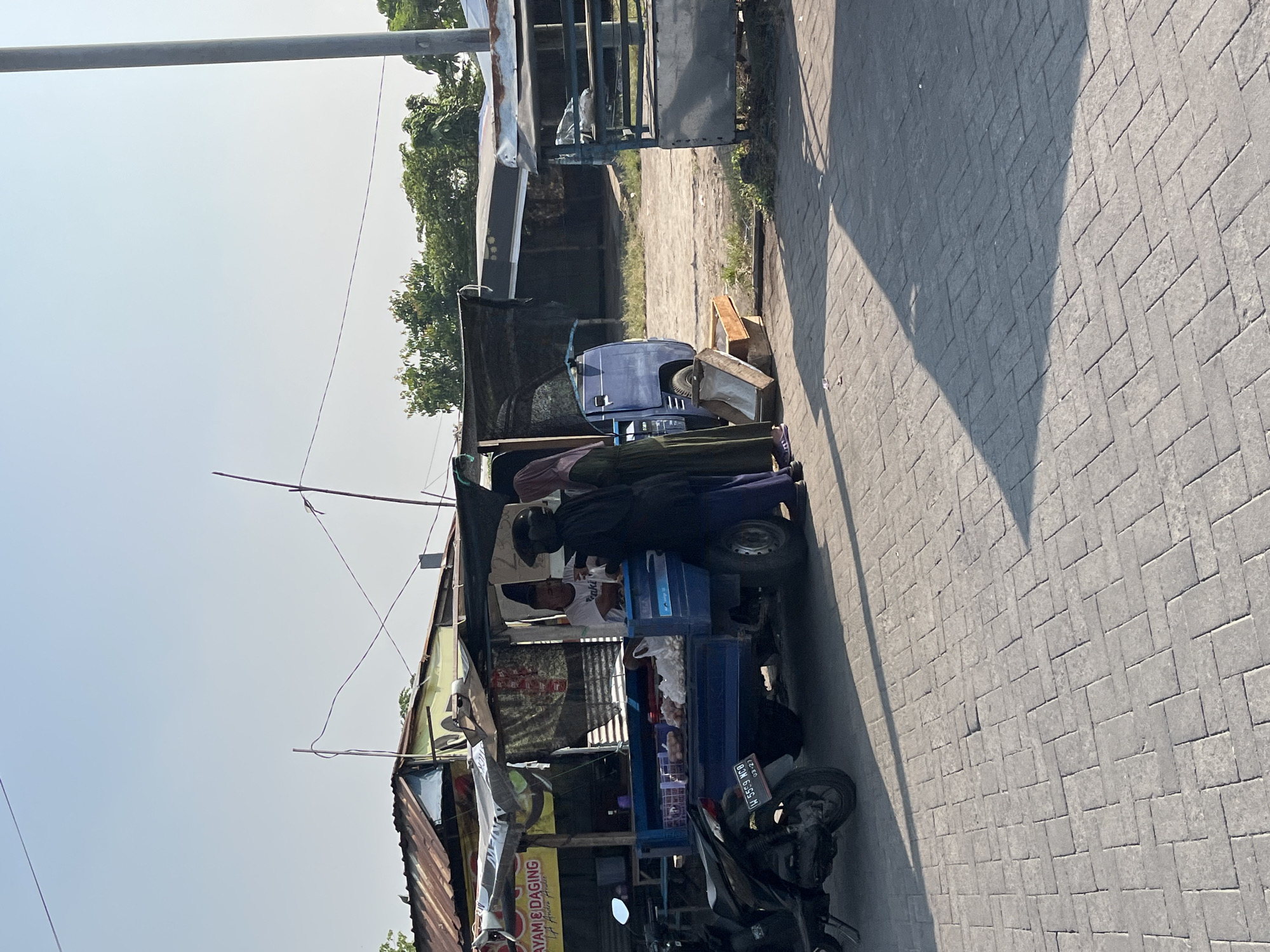 The image depicts a street scene in Kesambi, Indonesia, on a clear day. The primary focus is a blue pickup truck, modified into a food stall, selling what appears to be meat products, indicated by a sign reading "AYAM & DAGING". The stall is shaded by a dark tarp. Three individuals are present at the stall: a man in a white shirt, presumably the vendor, is interacting with two women. One woman, wearing a hijab and a dark outer garment, is facing the vendor, while the other woman, also in a hijab and a long olive-green dress, stands slightly behind her. A black motorcycle is parked to the left of the truck. The foreground shows a paved walkway with a brick pattern, and shadows are cast by the stall and the motorcycle. In the background, other simple structures with corrugated tin roofs and trees are visible, suggesting a roadside market or an informal commercial area. The overall atmosphere is that of an active marketplace under a bright sky.