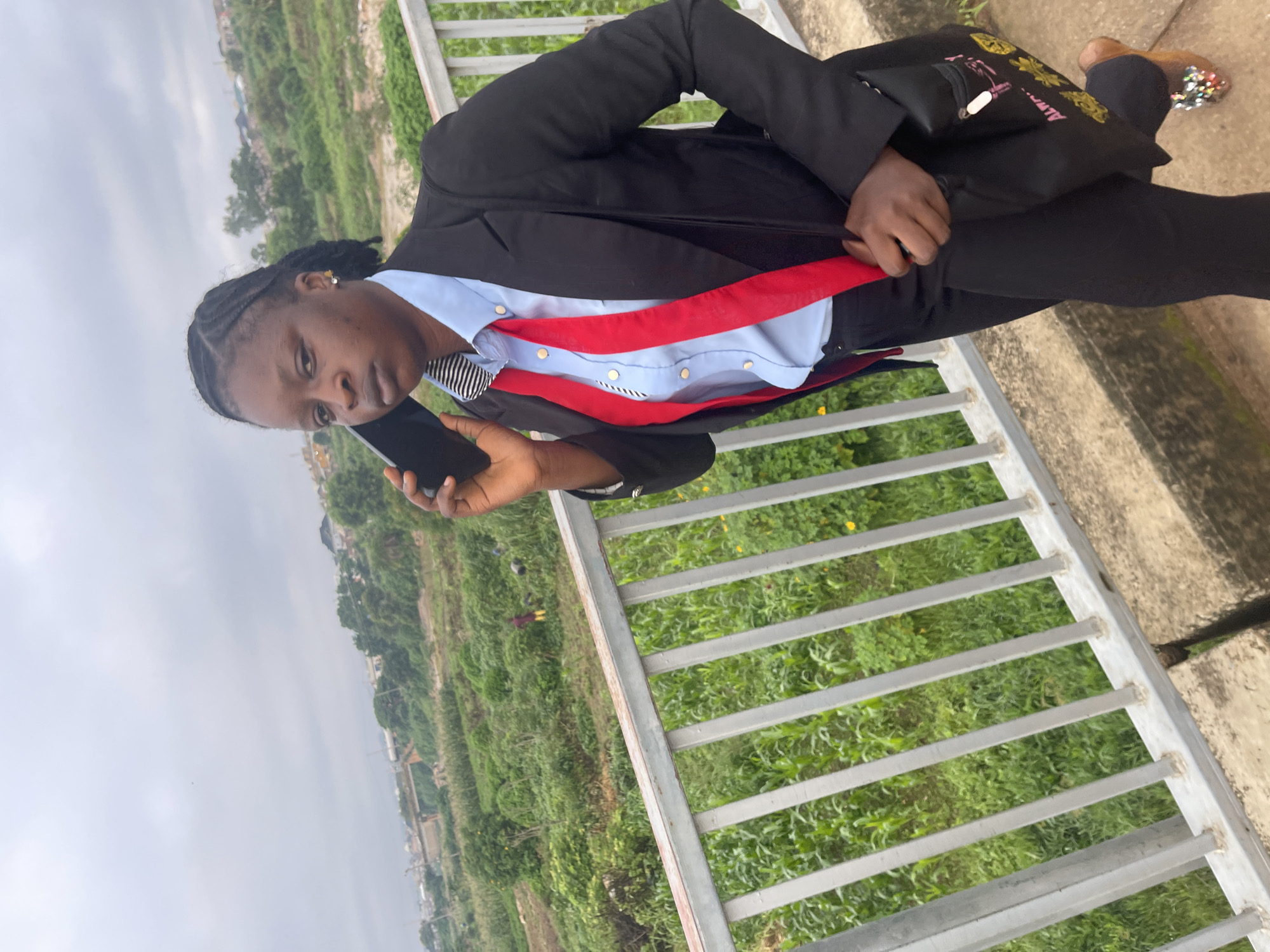 A young Black woman wearing a black suit, light blue dress shirt, and a red tie, stands on a concrete walkway with a metal railing. She is holding a black smartphone to her left ear. The background shows a lush green landscape with scattered buildings and a cloudy sky. The image was taken in Rafin Par, Nigeria.