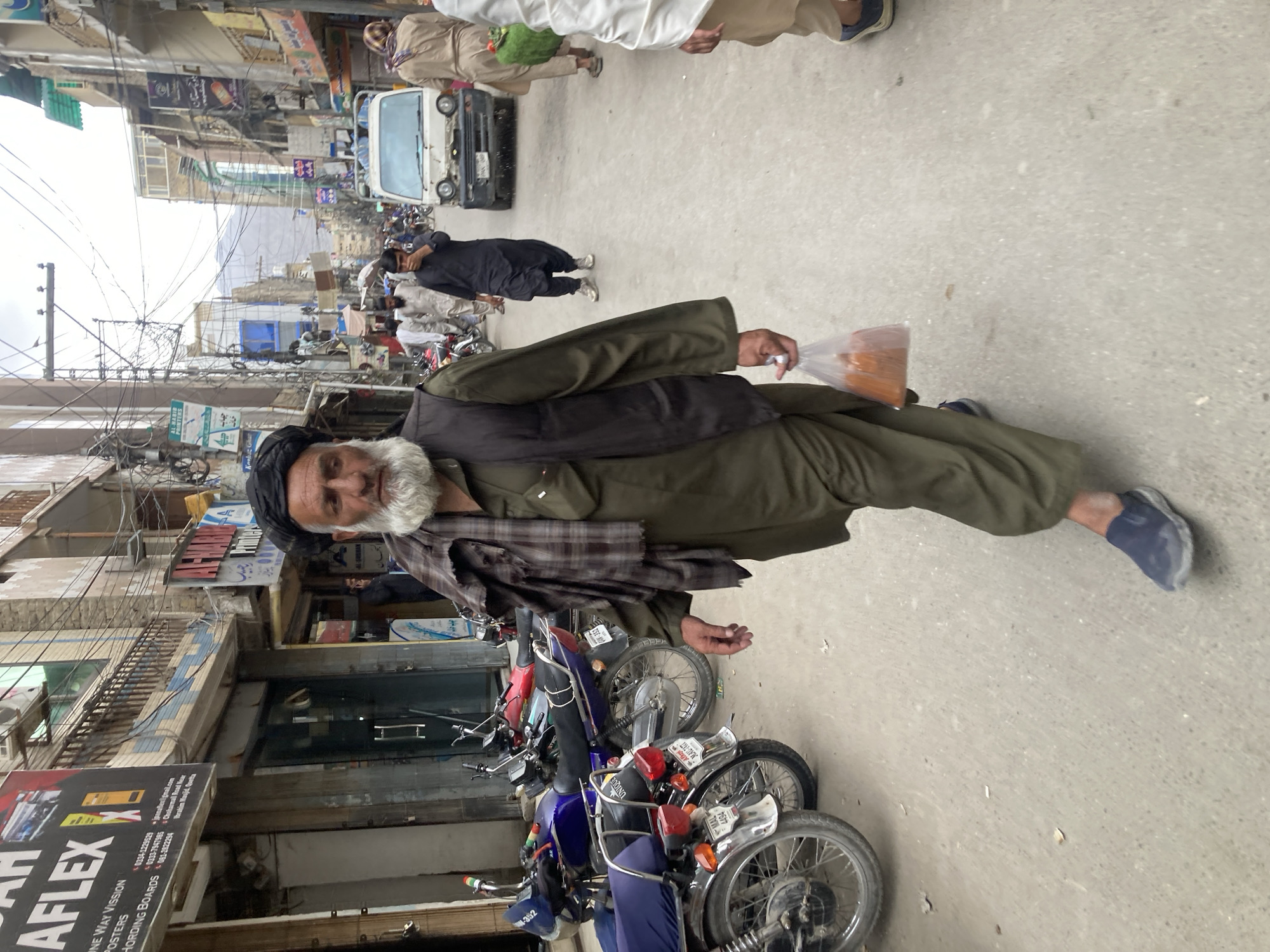 An elderly man with a white beard, wearing a black turban and green clothing, stands in the foreground of a street in Quetta City Tehsil, Pakistan. He holds a clear plastic bag filled with an orange-colored substance. Behind him, parked motorcycles are visible. A van and other pedestrians are further down the street. Buildings line the street, with various signs and advertisements. Overhead, power lines crisscross. A sign reading "AFLEX" is visible in the lower left corner.