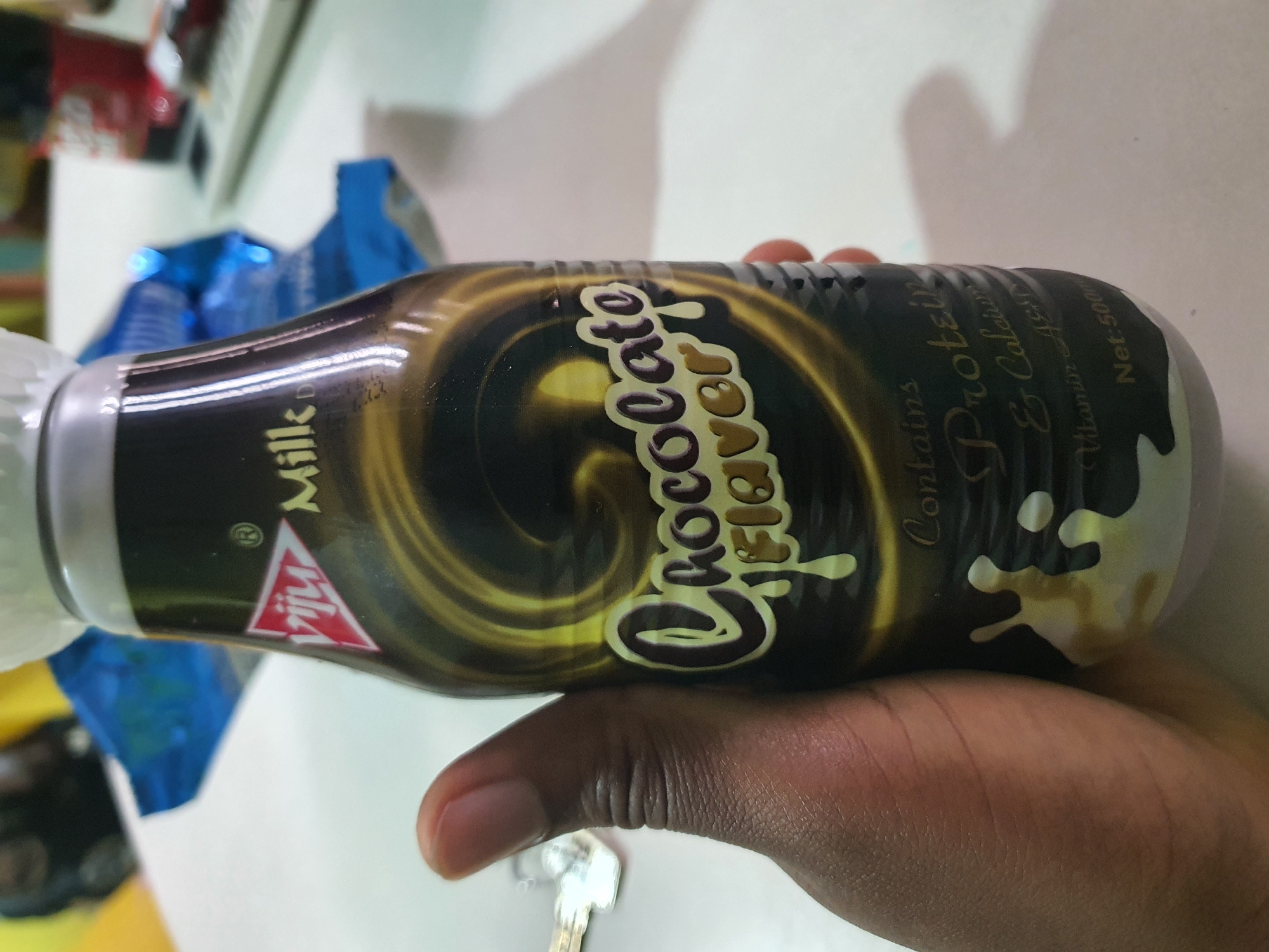 A person's hand, with dark skin, is holding a bottle of Viju Milk Chocolate Flavor drink. The bottle is dark brown with a swirling golden design and white text. The Viju logo, a red triangle with white text, is visible on the upper part of the bottle. The background is blurred and appears to be an indoor setting with a white table and some blue and red objects in the distance, suggesting it could be a home or office environment in Palmgrove, Nigeria. The lighting is natural and casts subtle shadows.